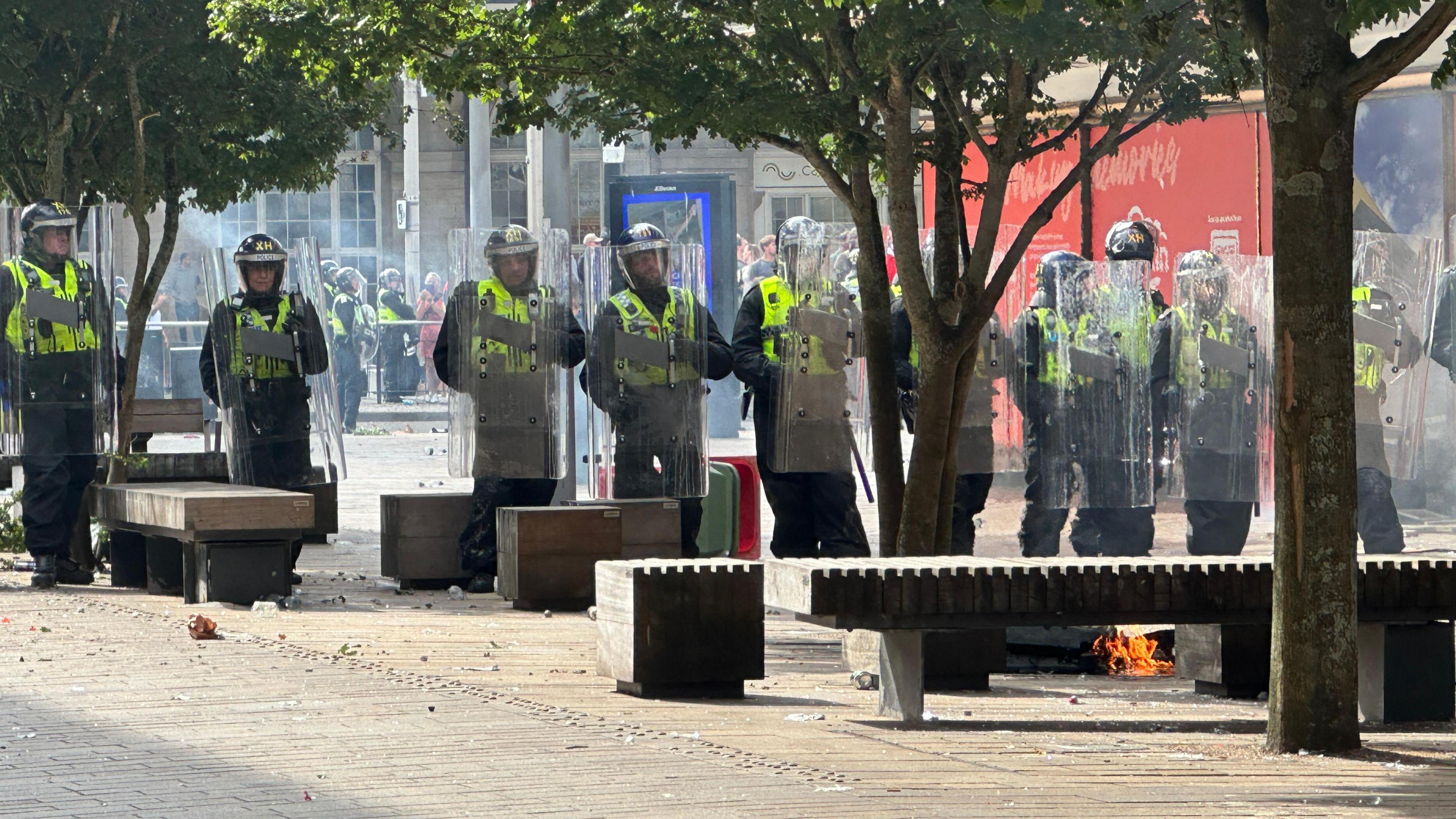 A line of police officers wearing riot gear and holding shields stand side by side, surrounded by benches and trees