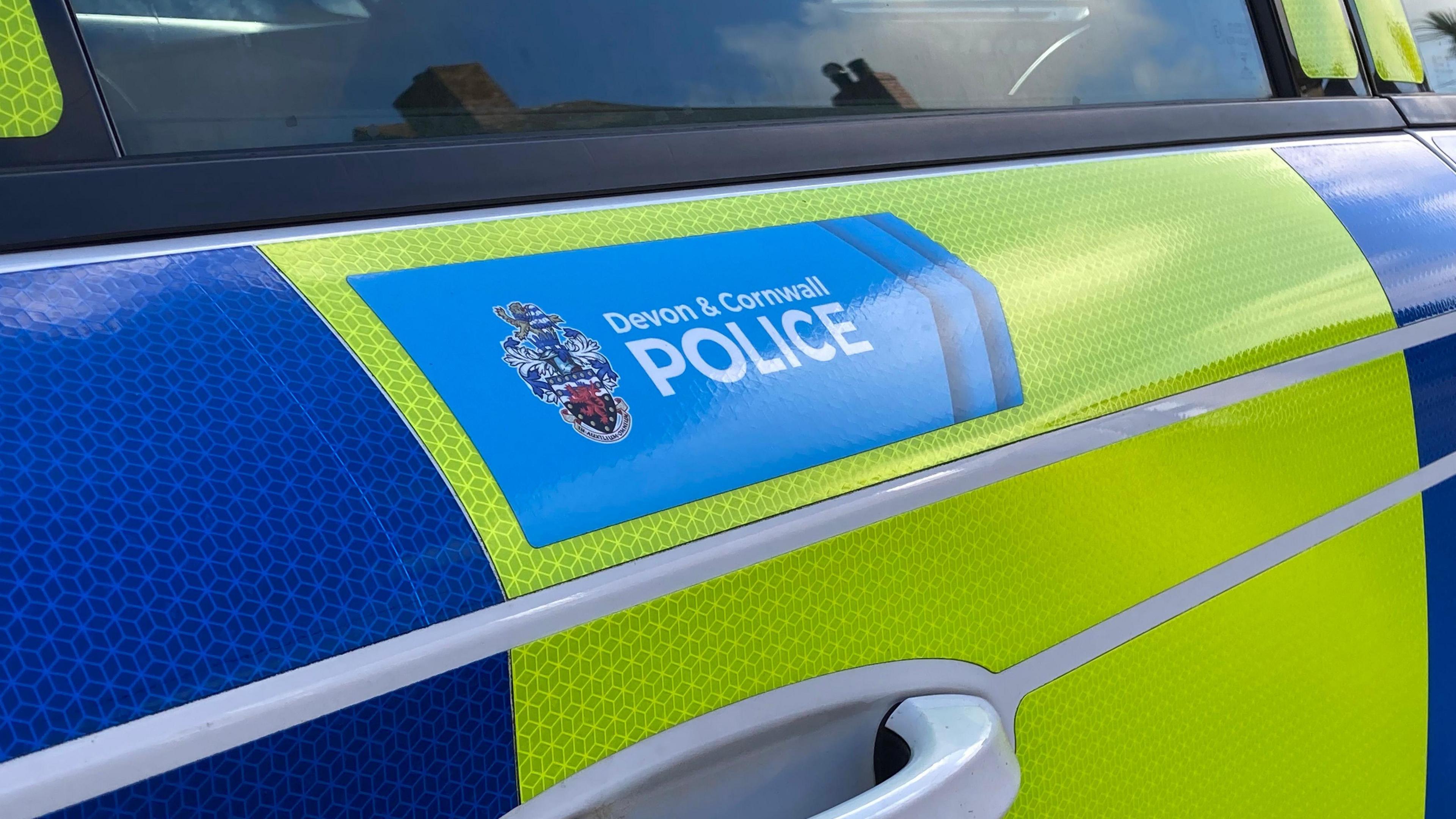 A close up shot of the outside door of a Devon & Cornwall Police car. The forces branding is on the outside in a blue rectangle. The regular police colours of yellow and blue are wrapped around the vehicle. 