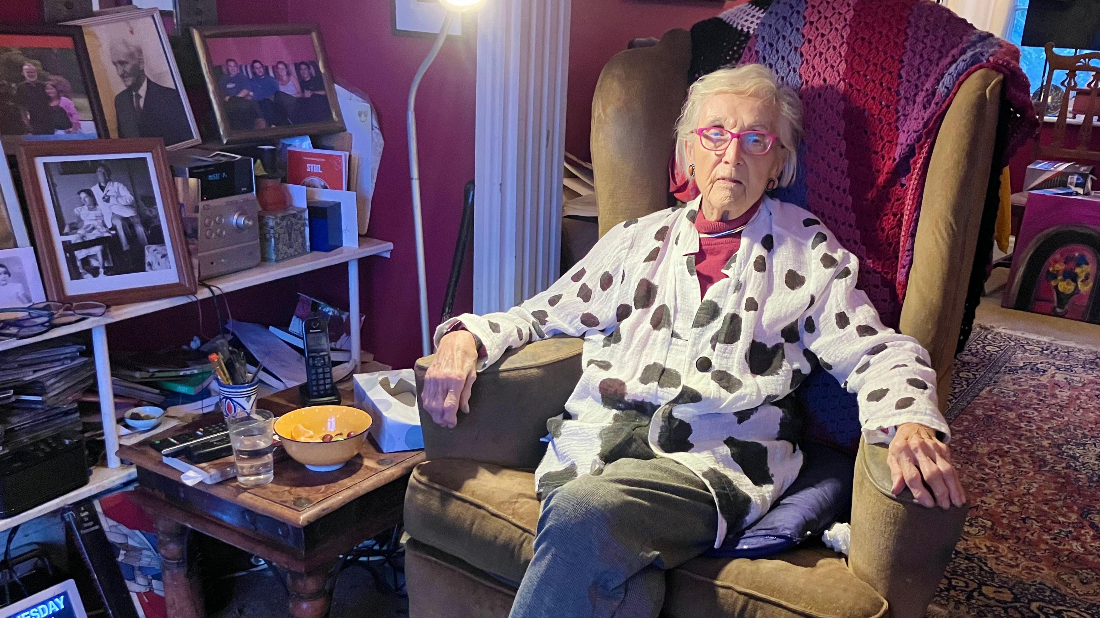 Glenys Cour sitting in a mustard armchair with a pink, red and purple blanket behind her. She has white short hair and is wearing pink glasses, pink earrings and a white blouse with black irregular spots. Besides her is shelving full of family photographs.