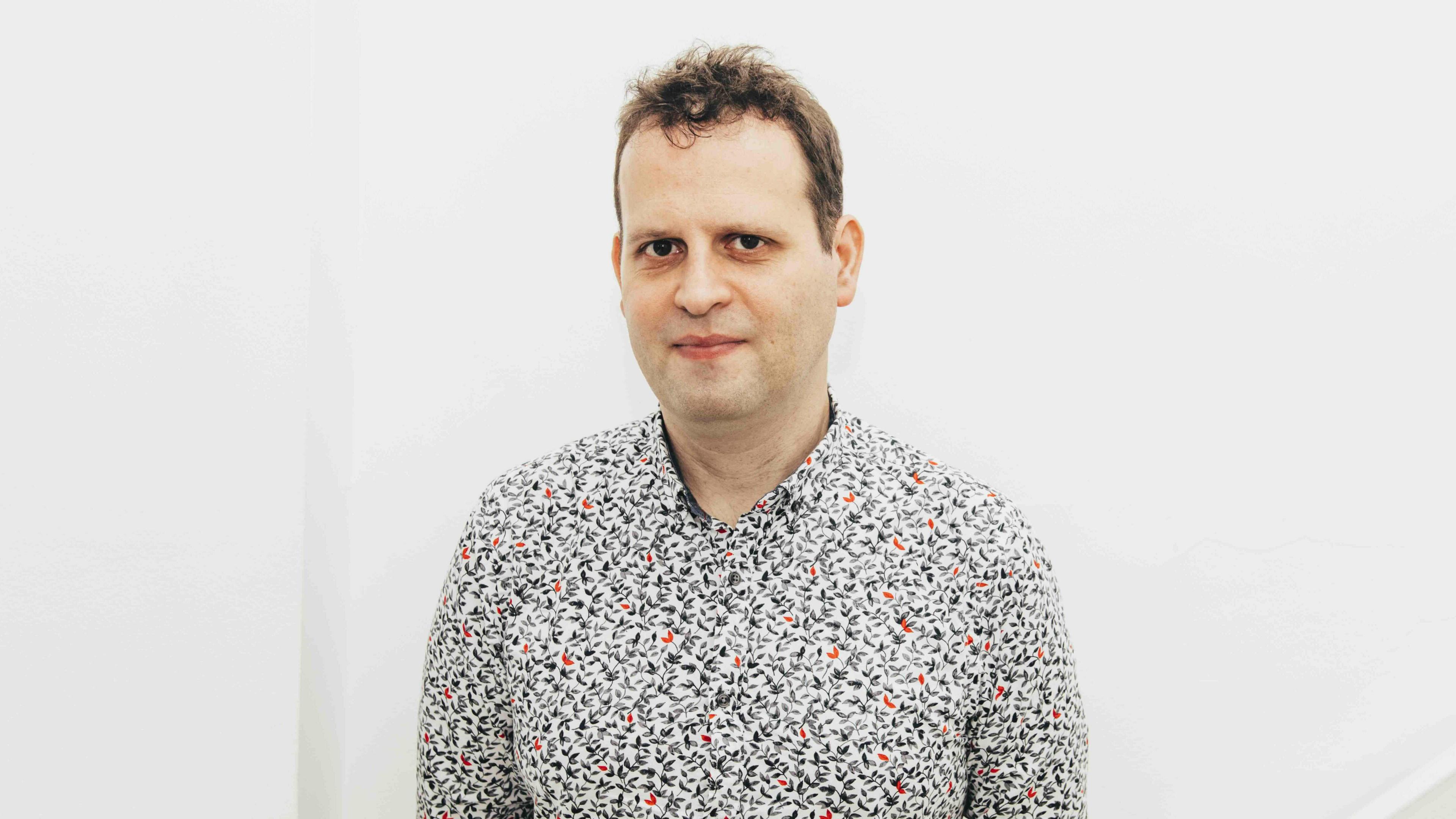 Adam Kay is a white man with short brown hair which is wispy and wavy on the top of his head. He is standing in front of a white wall and wearing a white shirt with a black and red patter on it.