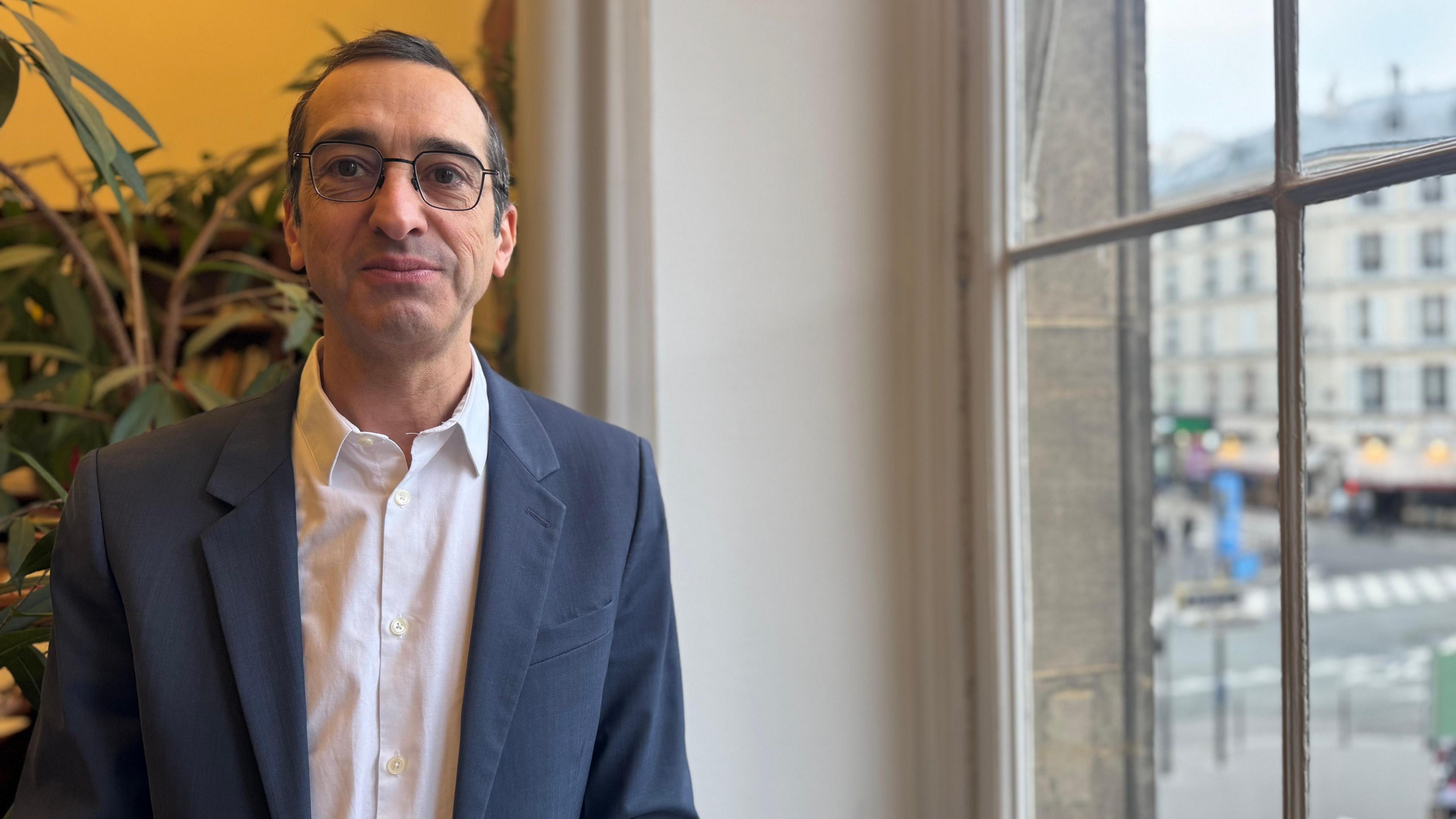 Rémi Féraud, a white man with glasses, wearing a white shirt and blazer, stands next to a window and in front of a yellow wall and green plant