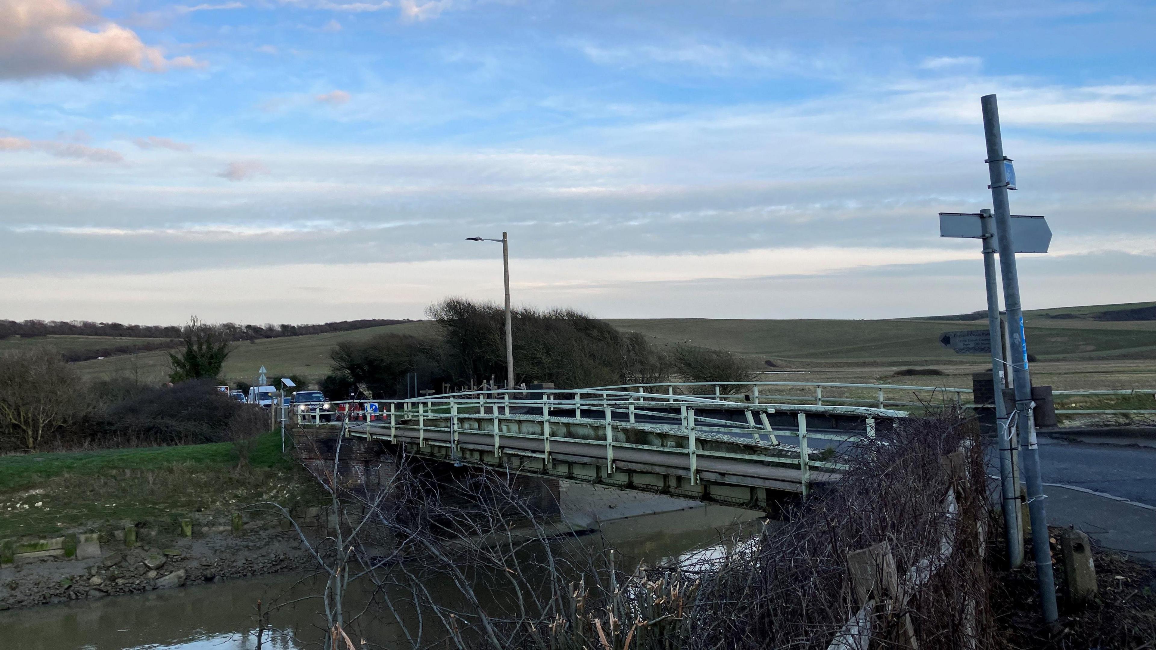 The single lane bridge with the river running underneath it. Cars can be seen on the far side and in the distance you can see the rolling hills of the South Downs.