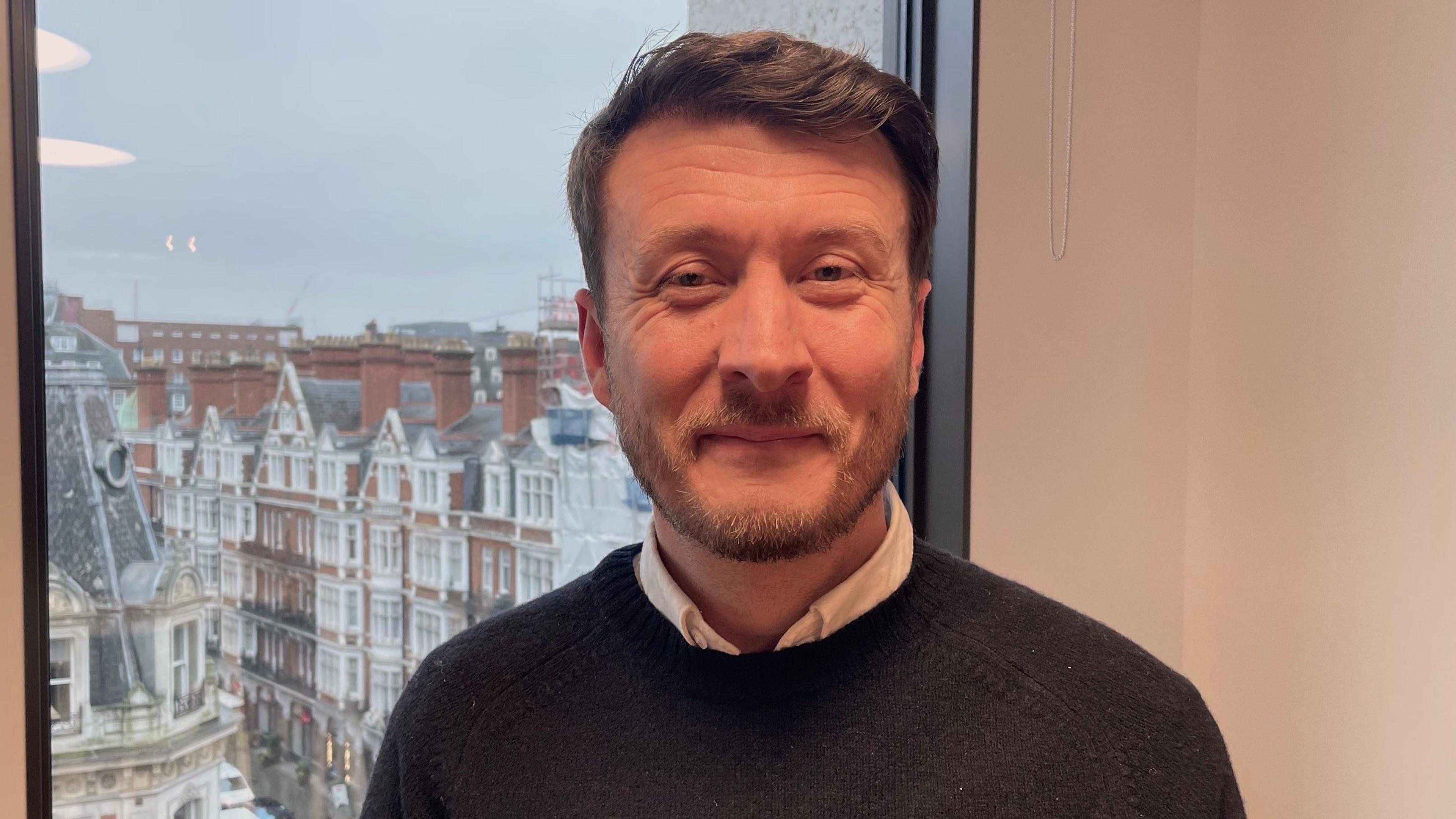 Michael Greslow looking direct to camera. He is smiling and wearing a dark coloured knitted jumper over a white shirt. He has a light coloured beard and short, brown hair brushed to one side. He is sat in front of a window which looks over an old-looking city centre, with five story red brick buildings with large white window frames.