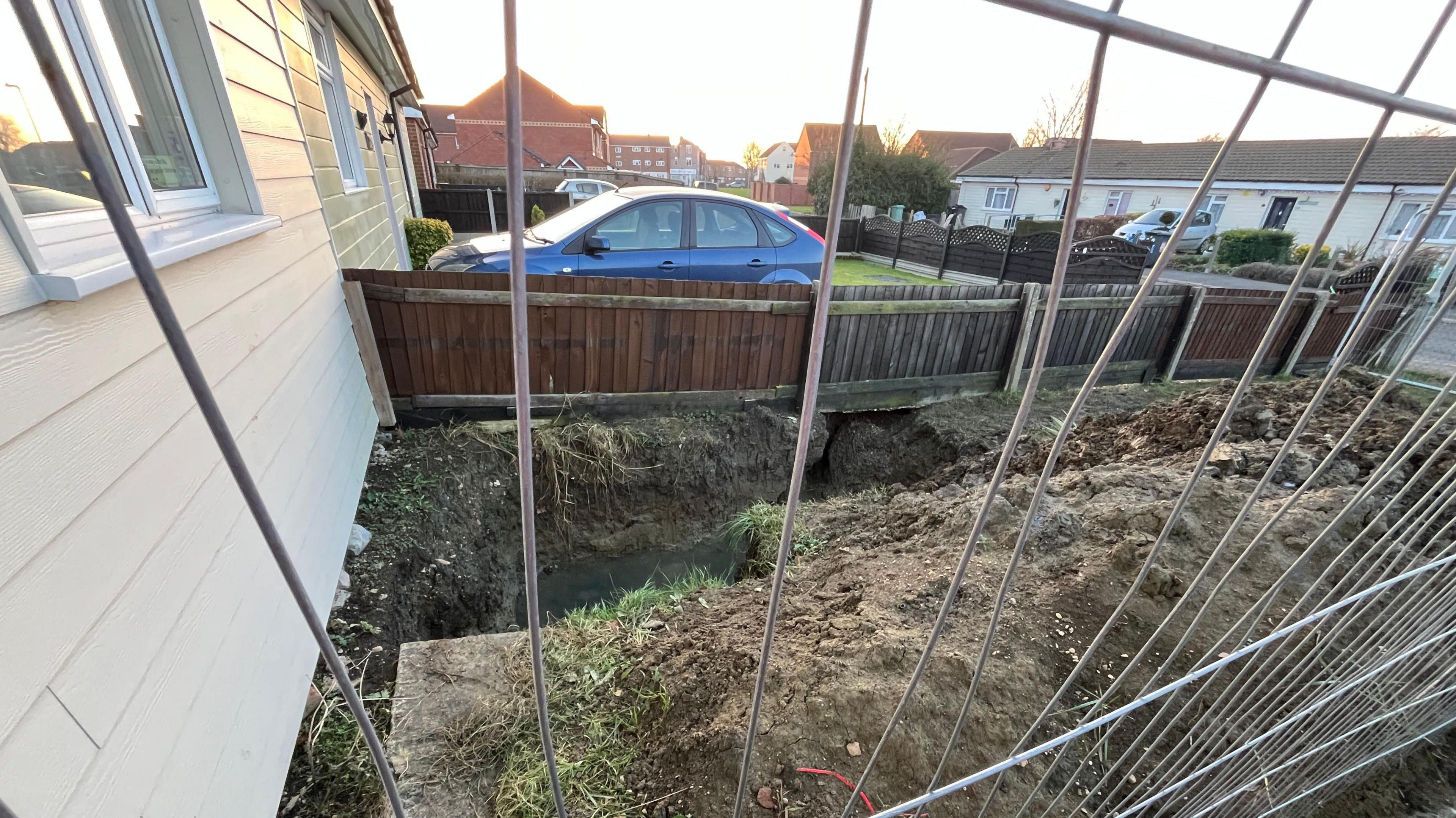 A view through a building fence on to a dug up trench filled with water. A part of it is opening towards the neighbour's driveway. 