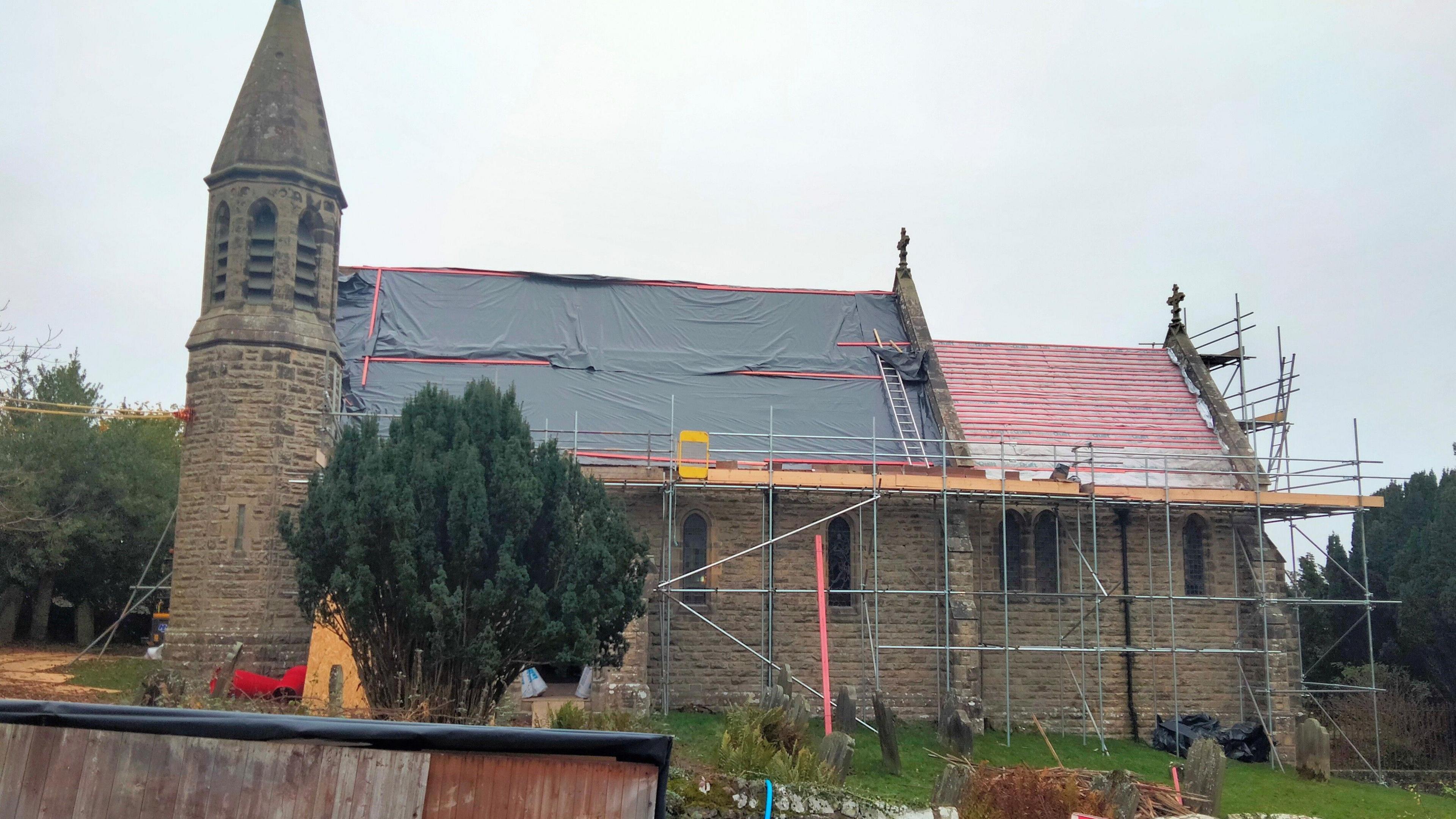 A Victorian village church surrounded by scaffolding with tarpaulin over the roof. 