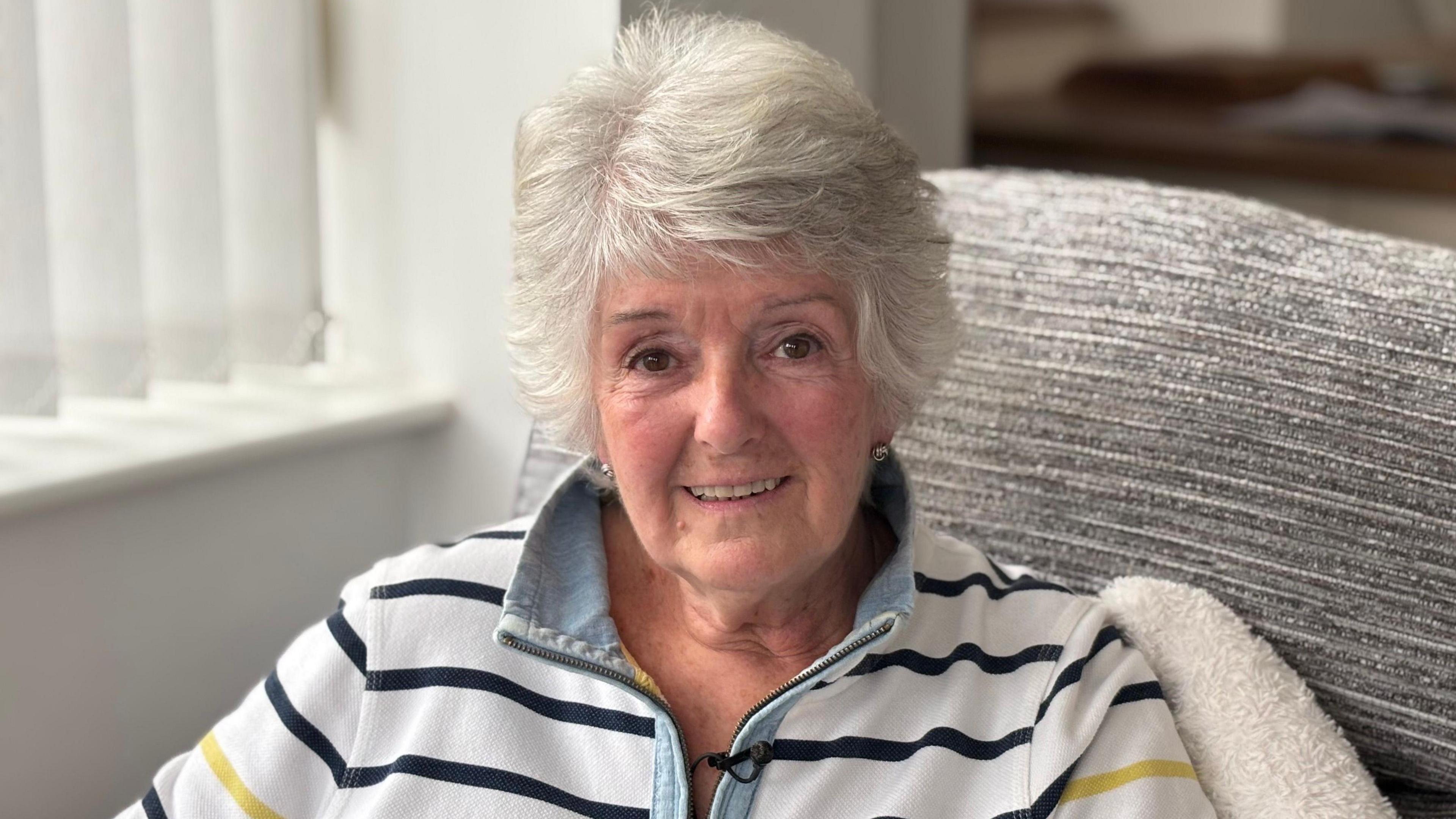 Joan Catherall yn gwenu at y camera wrth eistedd yn sedd lwyd yn ei thŷ. Mae'n gwisgo siwmper wen gyda streipiau glas tywyll. Mae ganddi wallt byr llwyd.