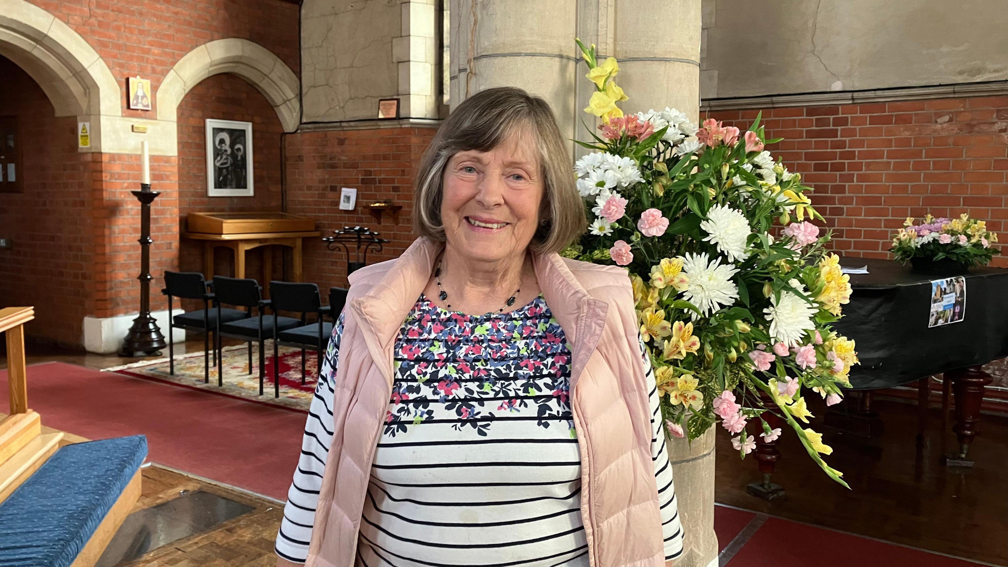 Jane Dobbie is wearing a striped top with flower patterns around the collar and is looking at the camera and smiling. She is standing in front of a bouquet of flowers in a church with a piano, chairs a stone walls in the background.