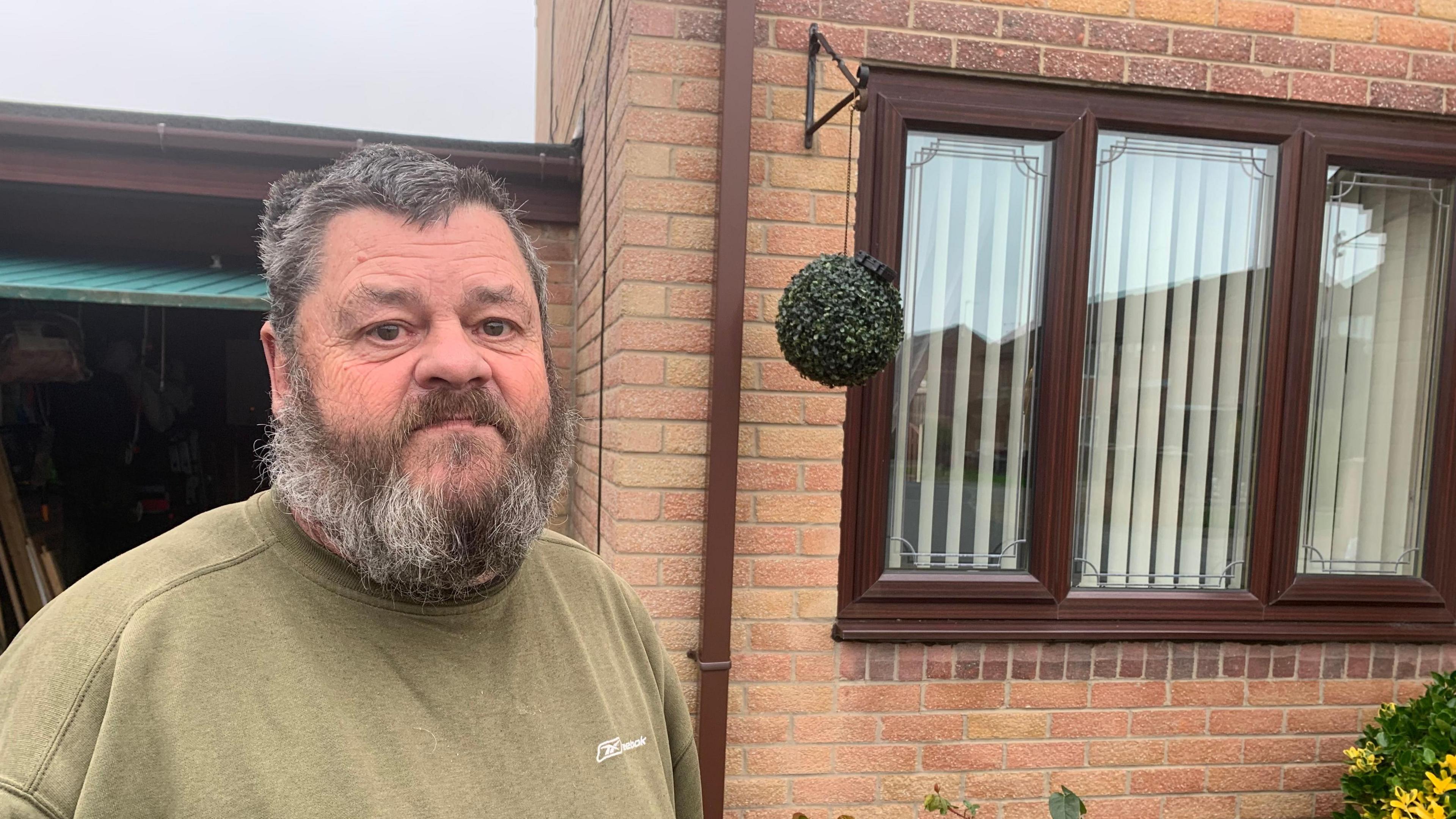 Peter Lewis has a big salt and pepper beard, short hair and a moustache. He is standing in front of his house in Catcliffe, Rotherham - a brick building with brown-framed PVC windows. He's wearing an olive green T-shirt.