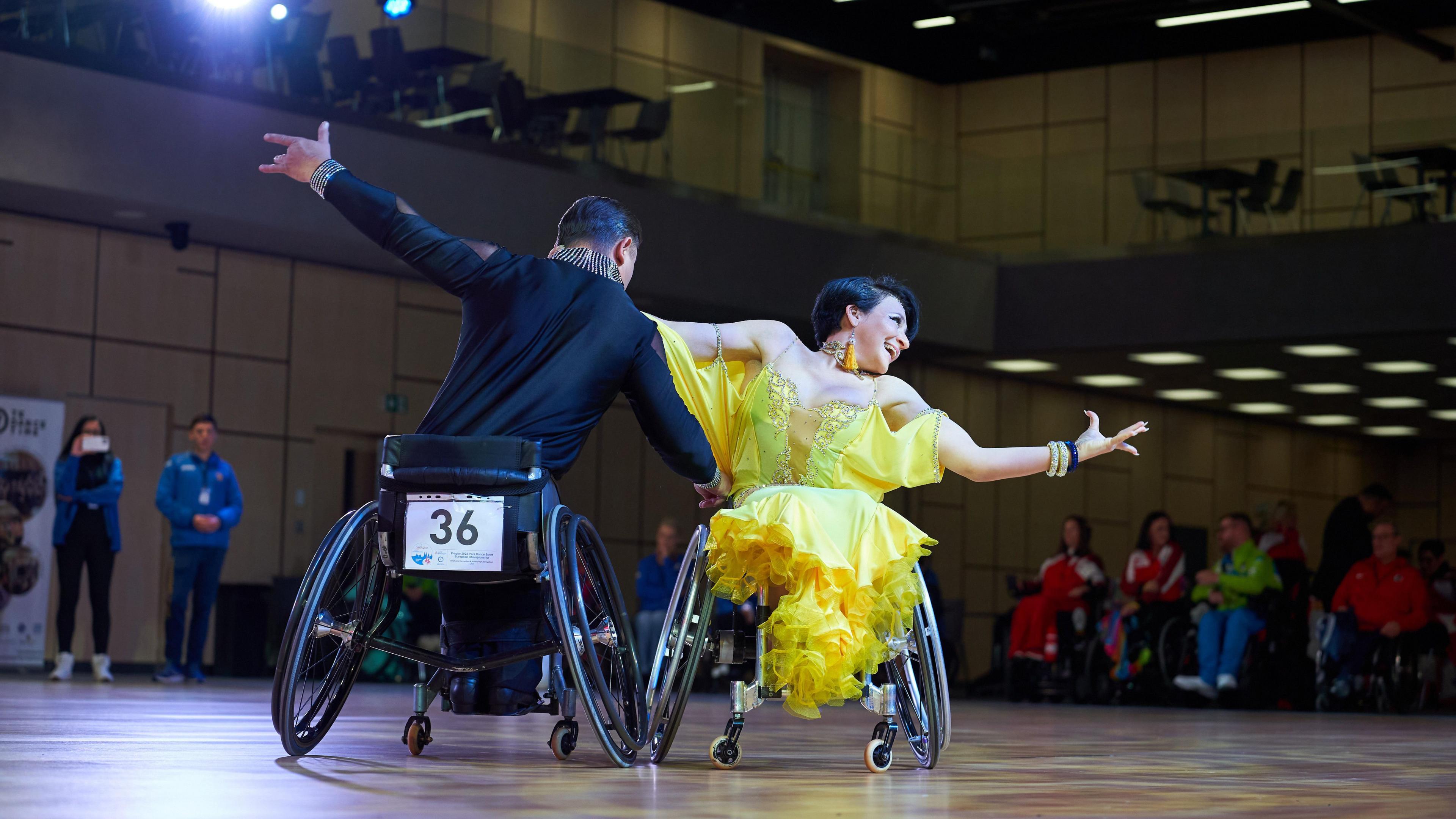 Two wheelchair users dance on a wooden floor with their arms outstretched. The lady has brown hair and is wearing a yellow ruffled dress, the man is wearing a black silk shirt with a bedazzled collar.
