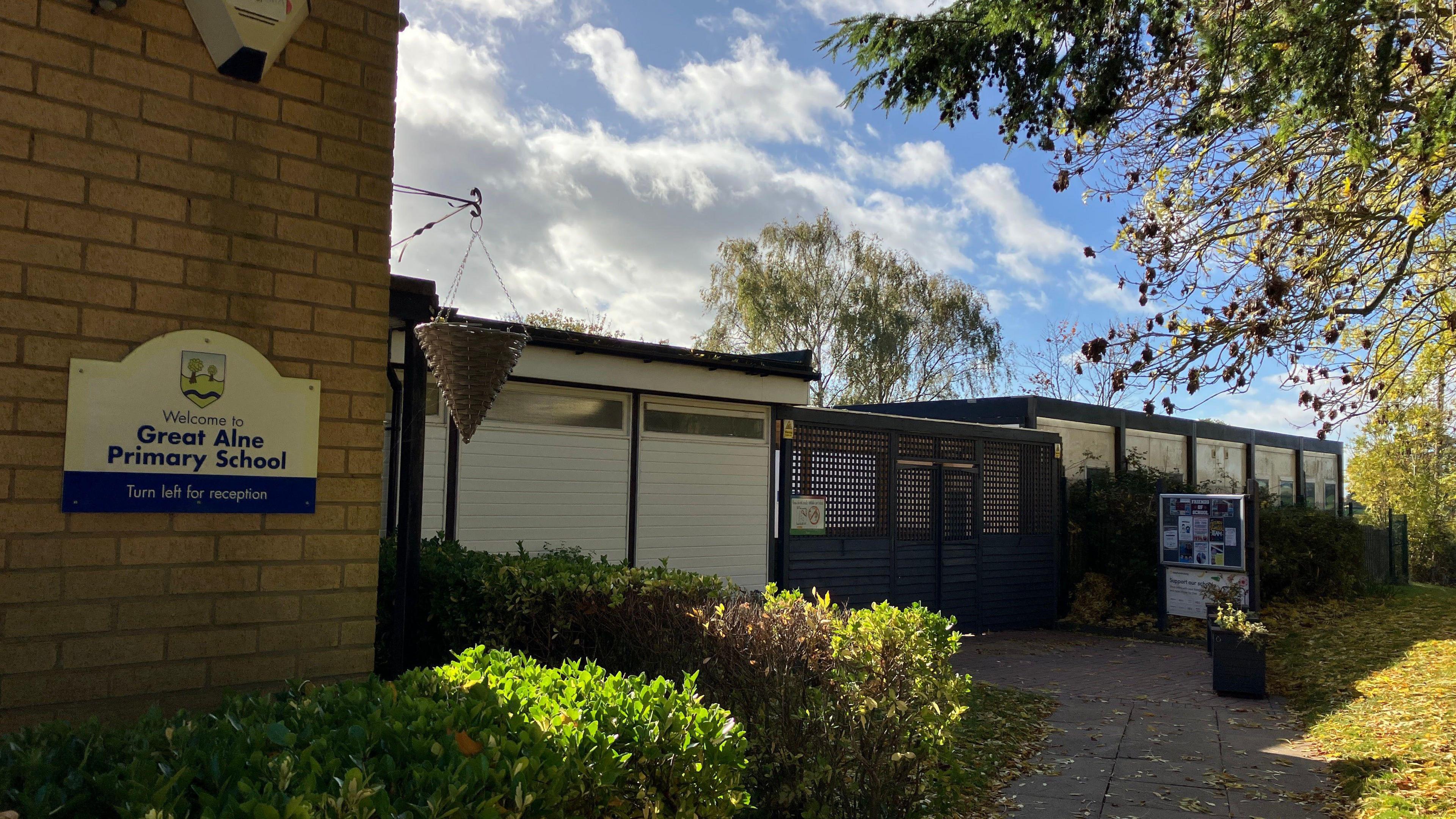 A school sign is fixed to a brick wall next to the school buildings. There are hedges, grassy areas and trees around the building. It is a sunny day.