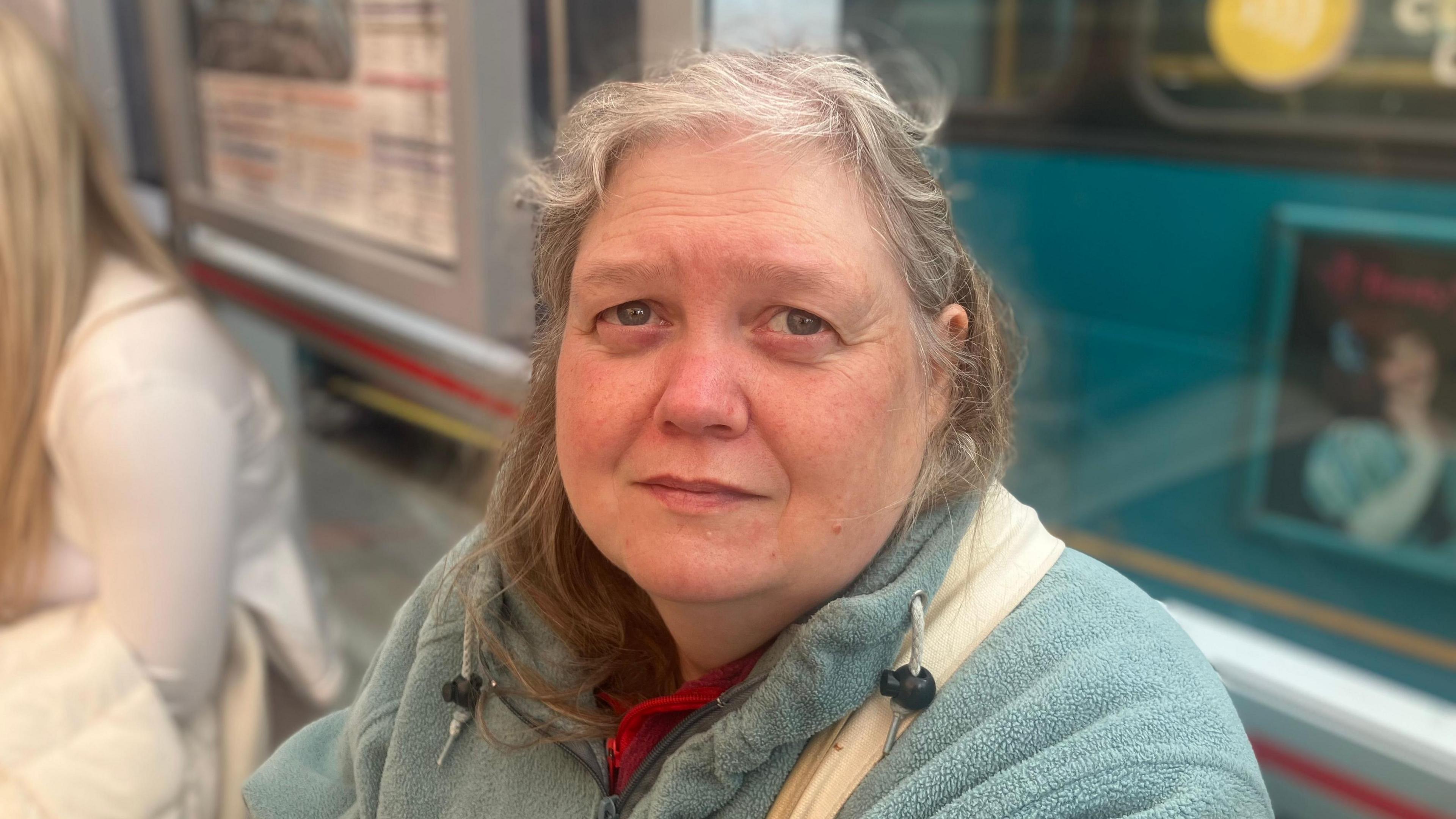 A woman with grey hair sits at a bus stop, wearing a blue fleece with a white bag over her shoulder. Behind her, a teal bus is blurred.
