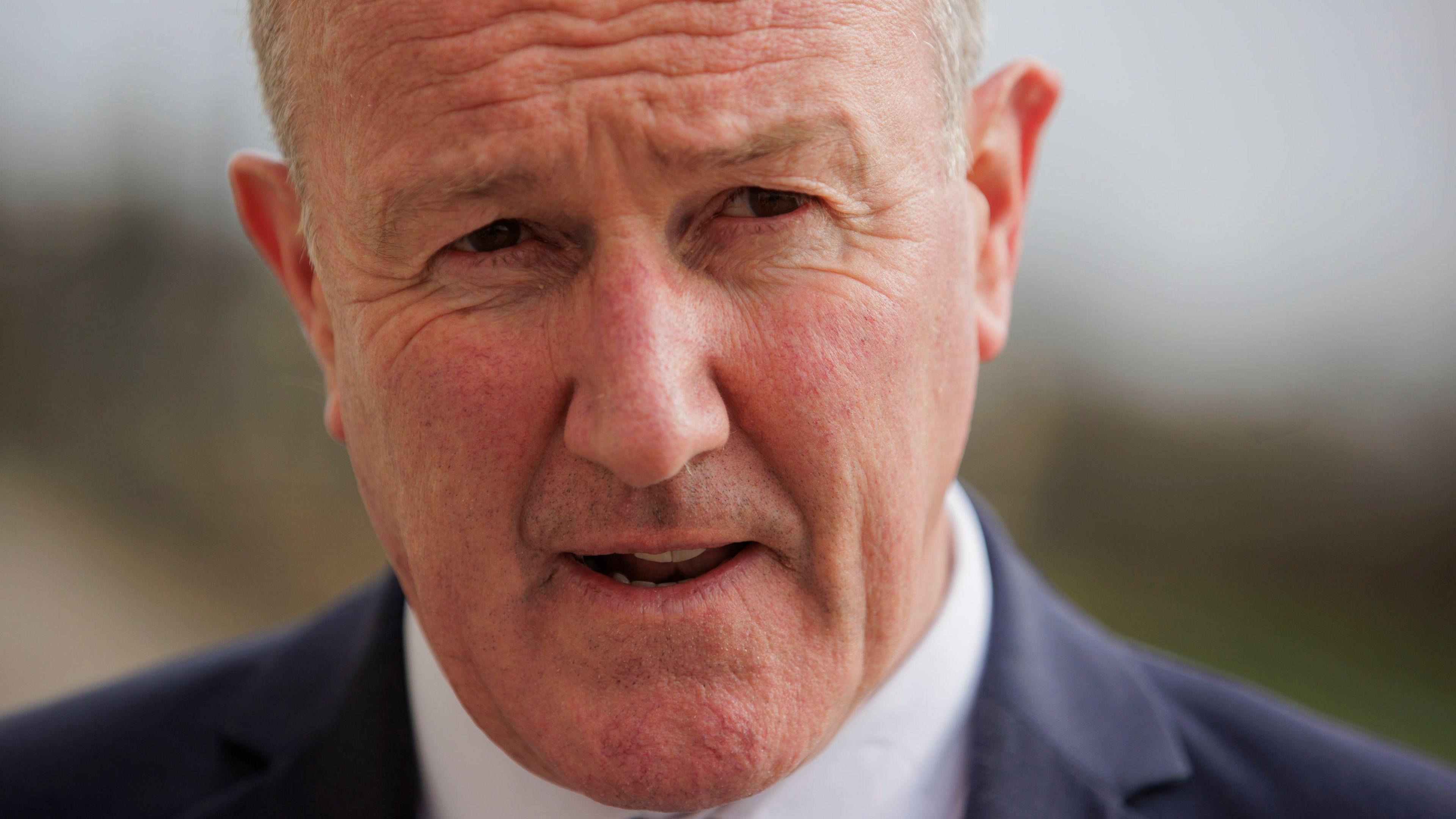 Conor Murphy at Parliament Buildings. He's wearing a dark coloured suit with a white shirt and a black and white checked tie. The photo is an extreme close up with the background blurred. He's speaking so his mouth is partially open.