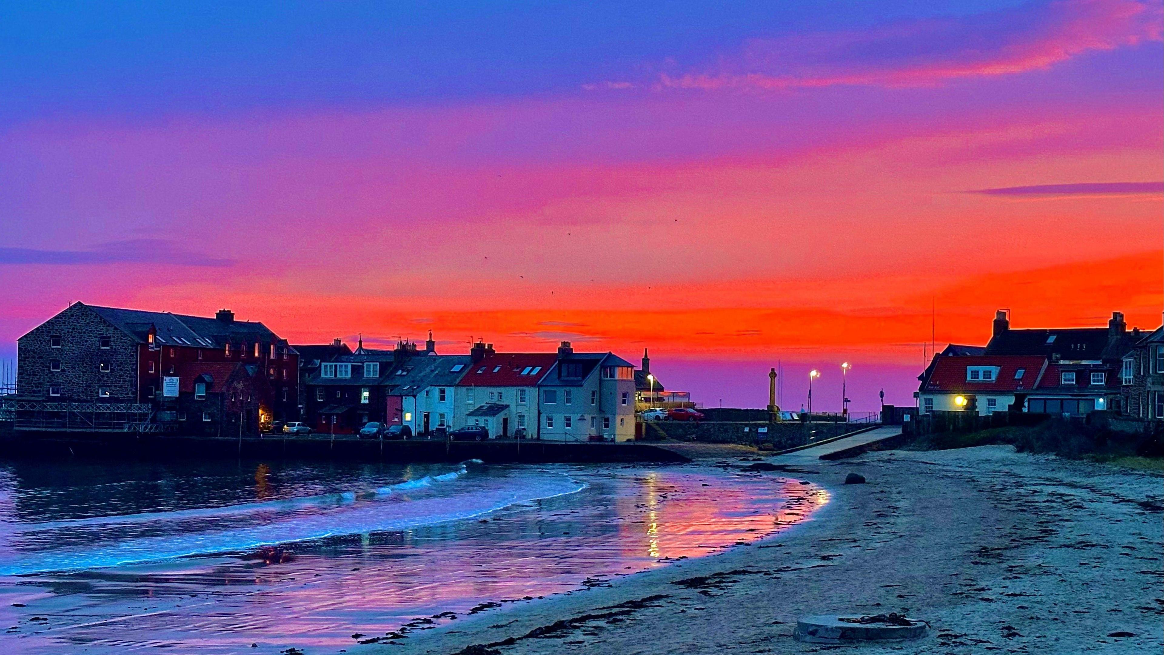 A blue, red and purple sky is the backdrop to a beach scene with houses at the far end.