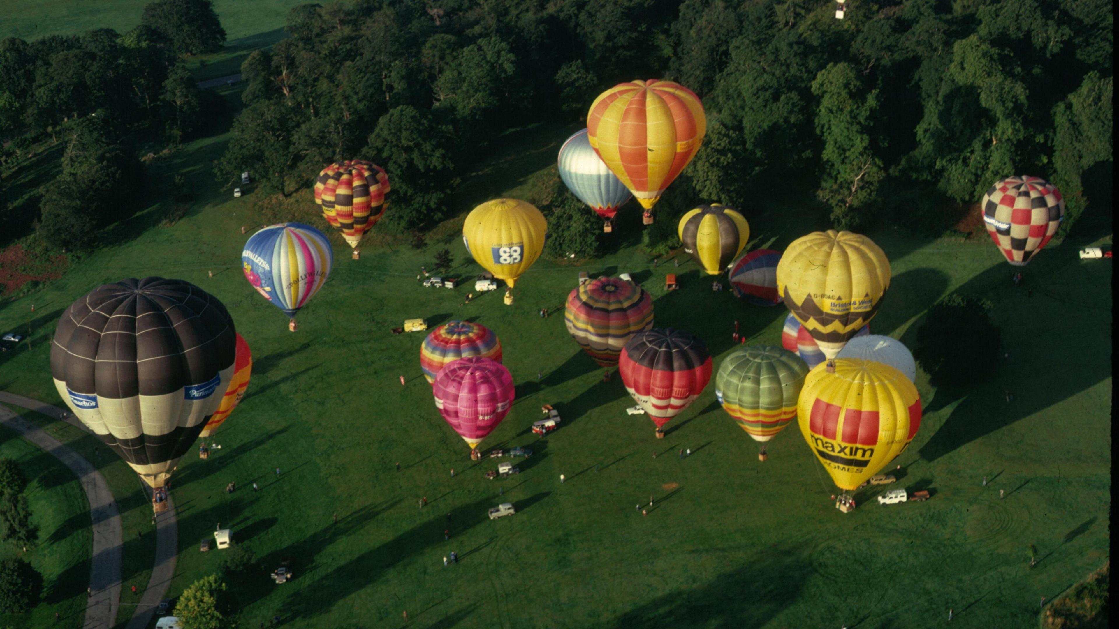 Various hot air balloons coloured yellow, green, red, pink, white and blue in different combinations flying as seen from a balloon at an even greater height