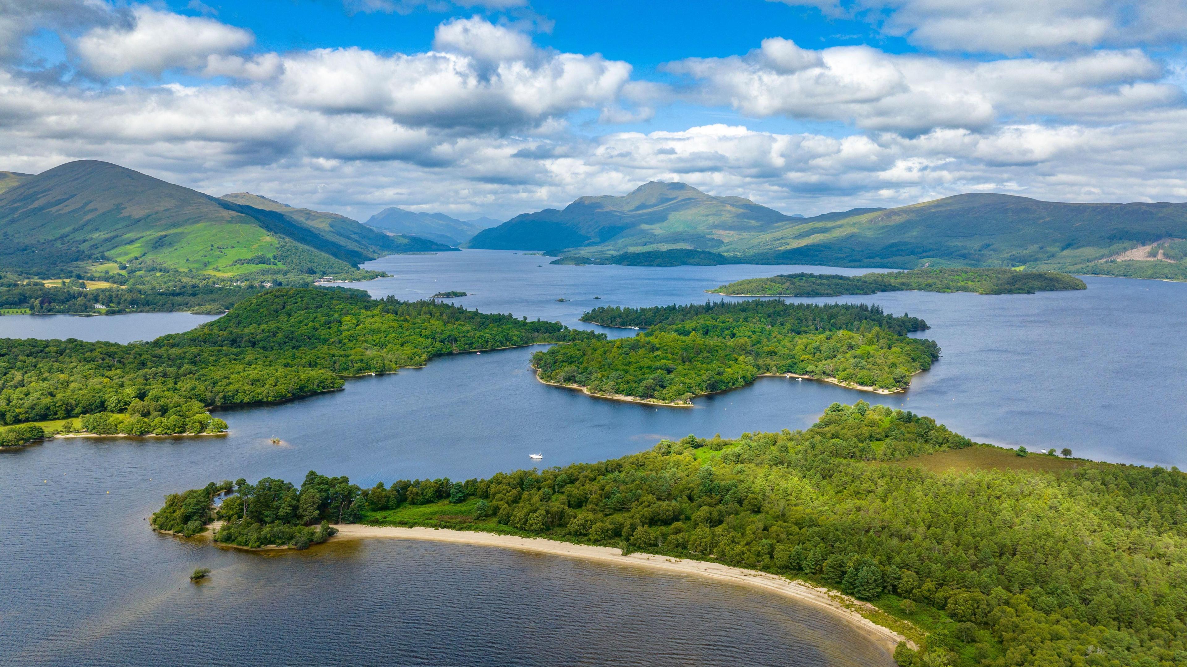 An aerial image of Loch Lomond, encompassing several of the lochs islands. The islands are beautiful shades of green and the water a rich blue. They lie in front of a rolling mountain range.