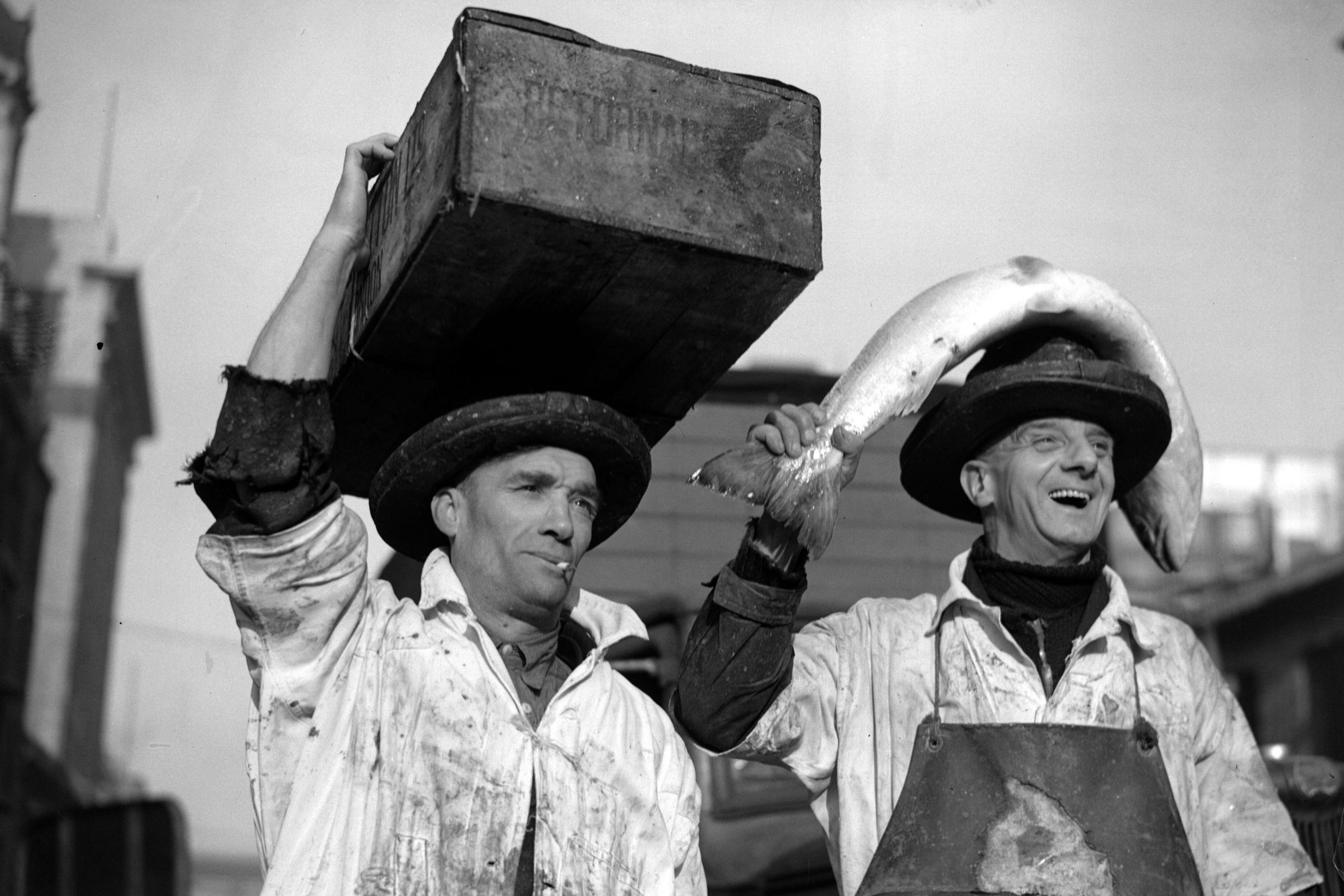 Black and white photo of two fish porters, one with a case on his head, the other with a large salmon on his head