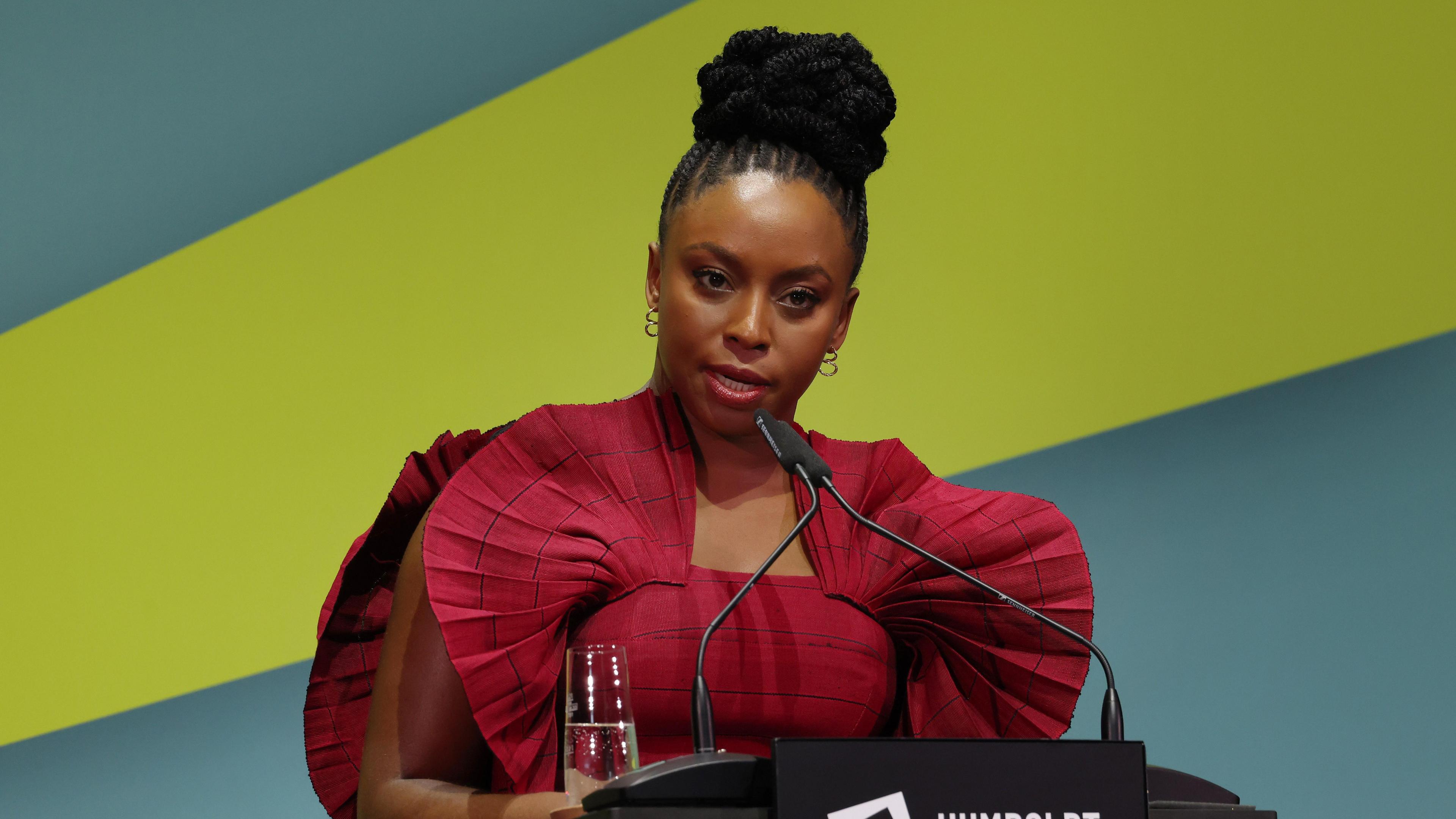 Chimamanda Ngozi Adichie, wearing a red shoulder fan dress, speaking into a microphone on a podium on stage, with a blue and green thick-striped wallpaper in her backdrop.