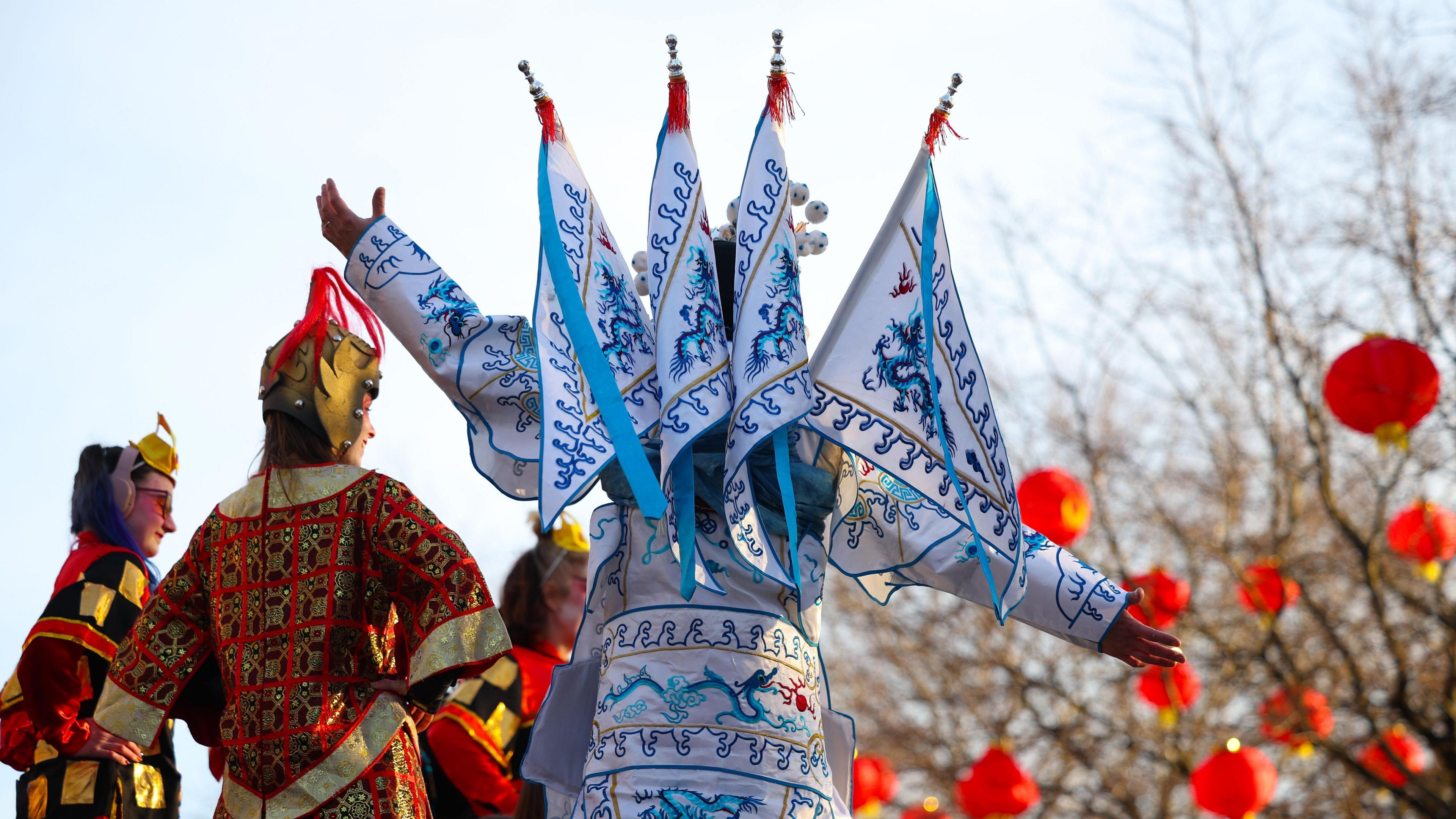 Stilt walkers in traditonal Chinese costumes, embroidered in intricate red and gold, and porcelain white and blue. Round red lanterns hang from tree branches in the background