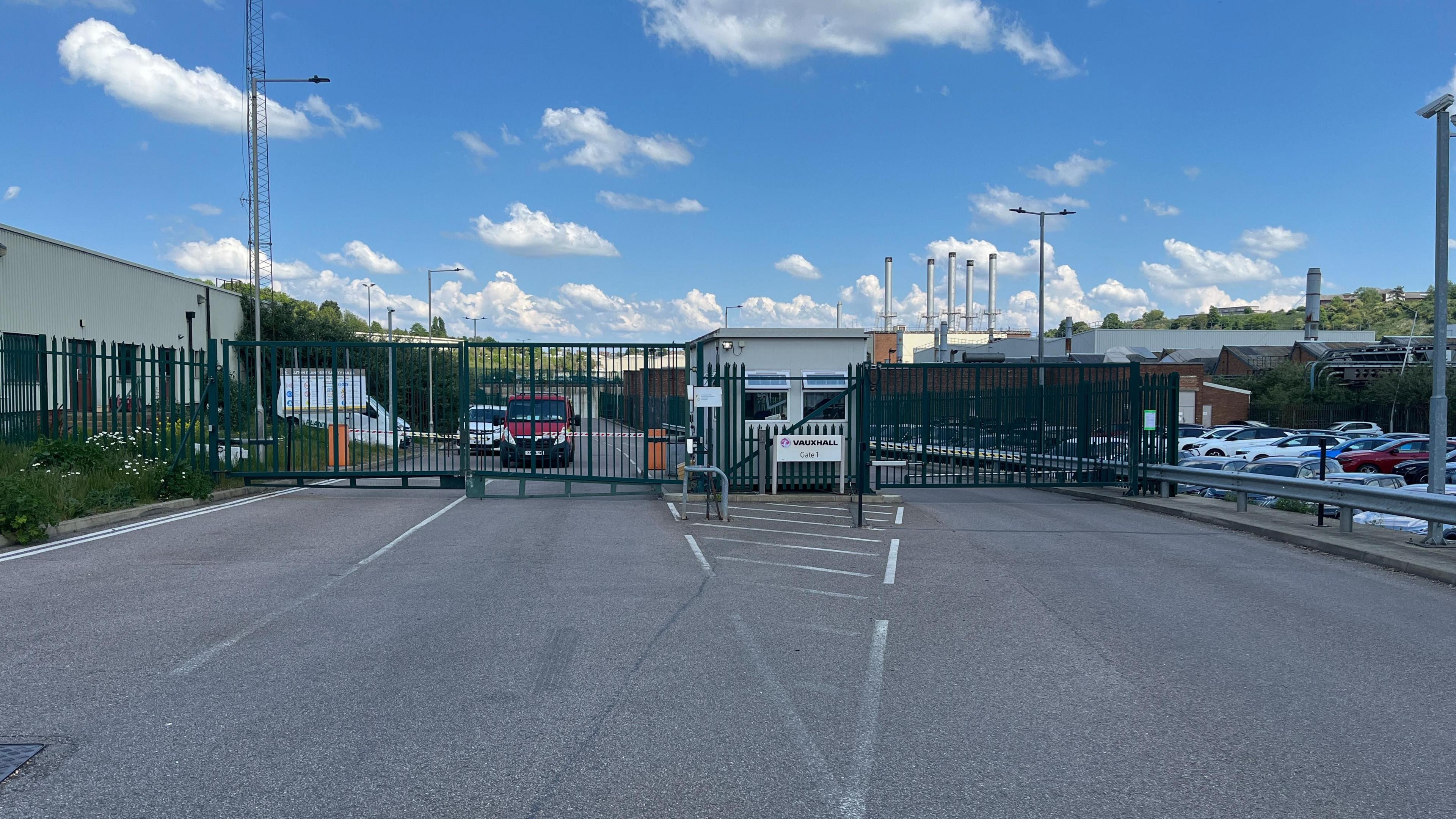A metal gate at the entrance to a Vauxhall factory with car parked in the background