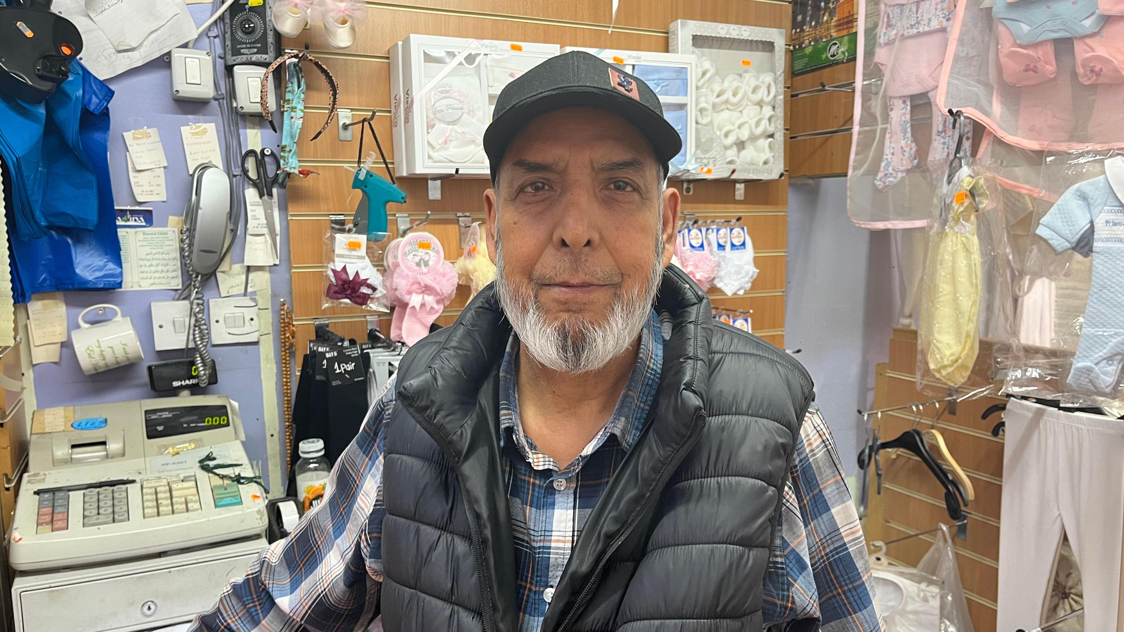 A man with a grey beard wearing a cap standing in front of a children's clothes shop
