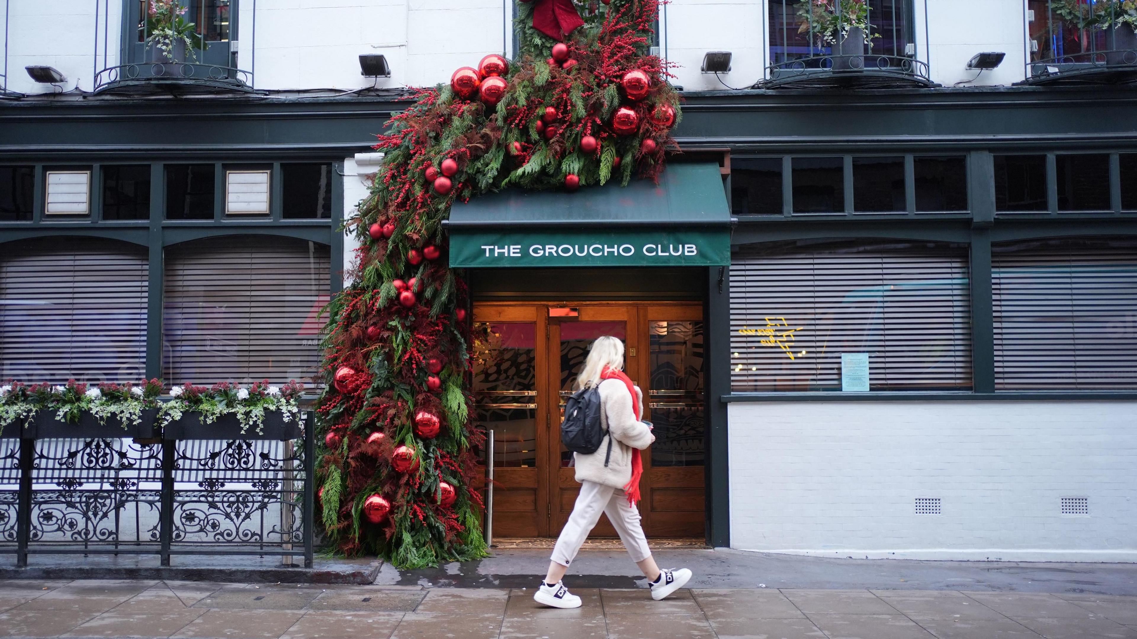 A woman is seen walking past The Groucho Club entrance. The wooden doors with windows have green awning with the club's name and it is decorated with a Christmas tree beside it