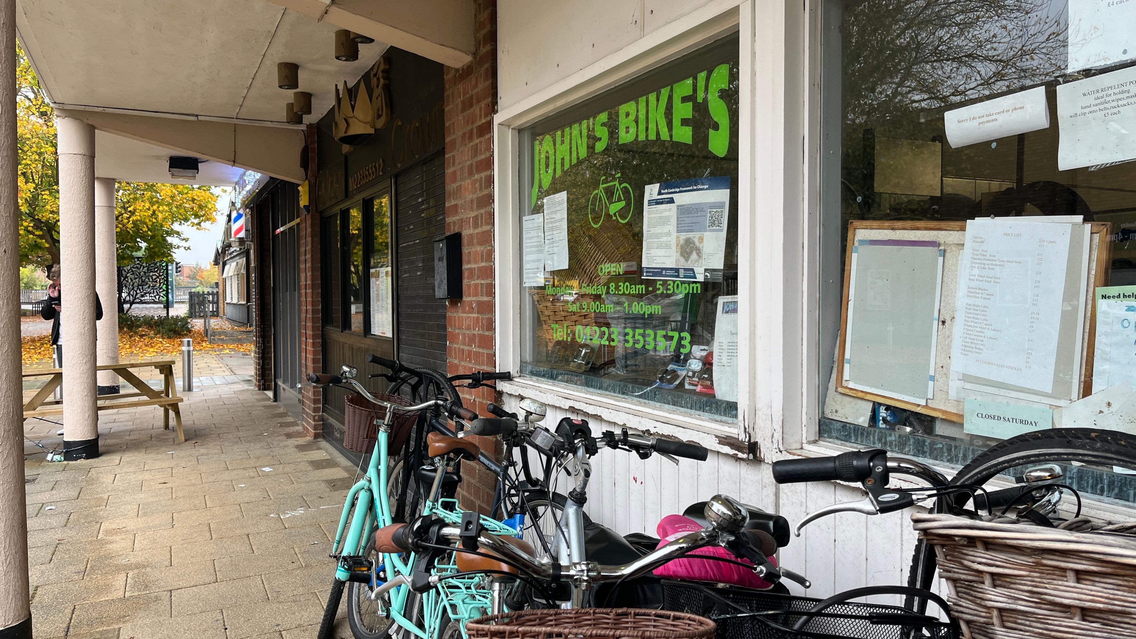 A bicycle shop with the name 'John's Bike's' prominently displayed in green lettering on the front window. Several bicycles, some with baskets, are parked outside on a paved walkway. The shop window features printed notices, including opening hours and a contact number. Autumn leaves are scattered nearby.