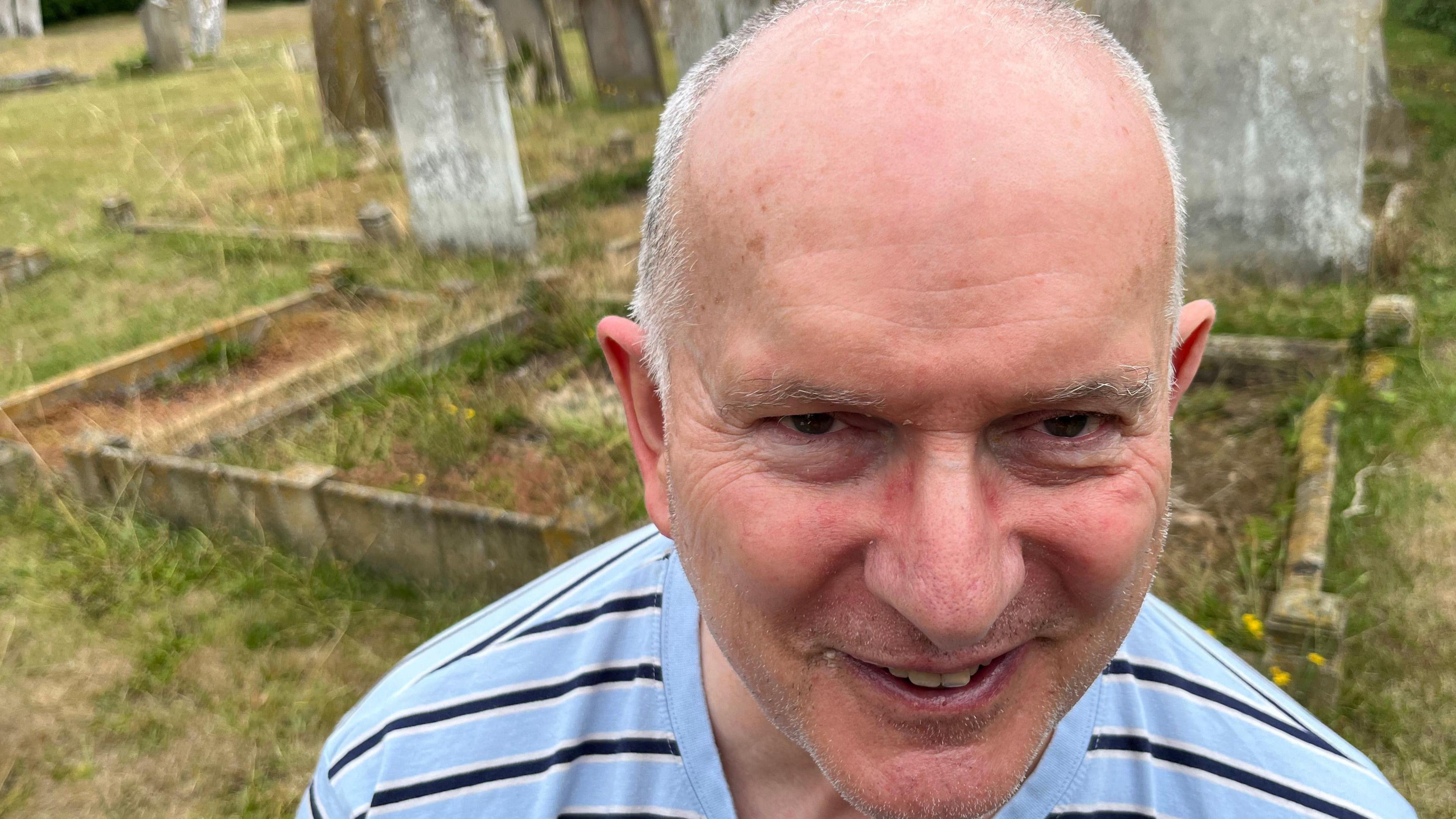 Mr Webb is smiling up at the camera. He bears some resemblance to his great uncle Mr Barker and they have similar mouth shapes. He is wearing a blue and black striped top and behind him are graves in the cemetery.