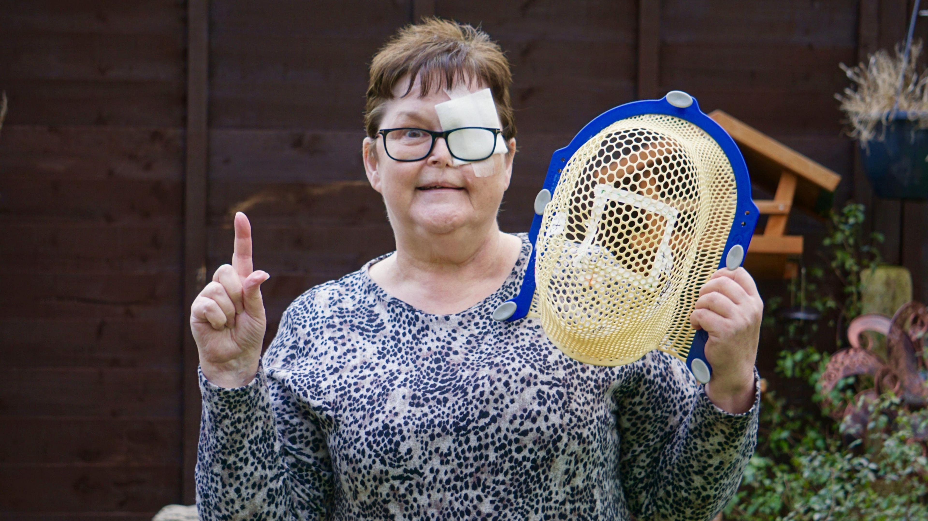 Melanie Wellings wearing a blue and white patterned long-sleeved top and black framed glasses. She has short brown hair and a white bandage over her left eye. She is standing in a garden in front of a brown fence, a wooden birdhouse, some bushes and hanging plants. She is holding a yellow and blue mesh hospital mask in one hand and holding her other index finger in the air.