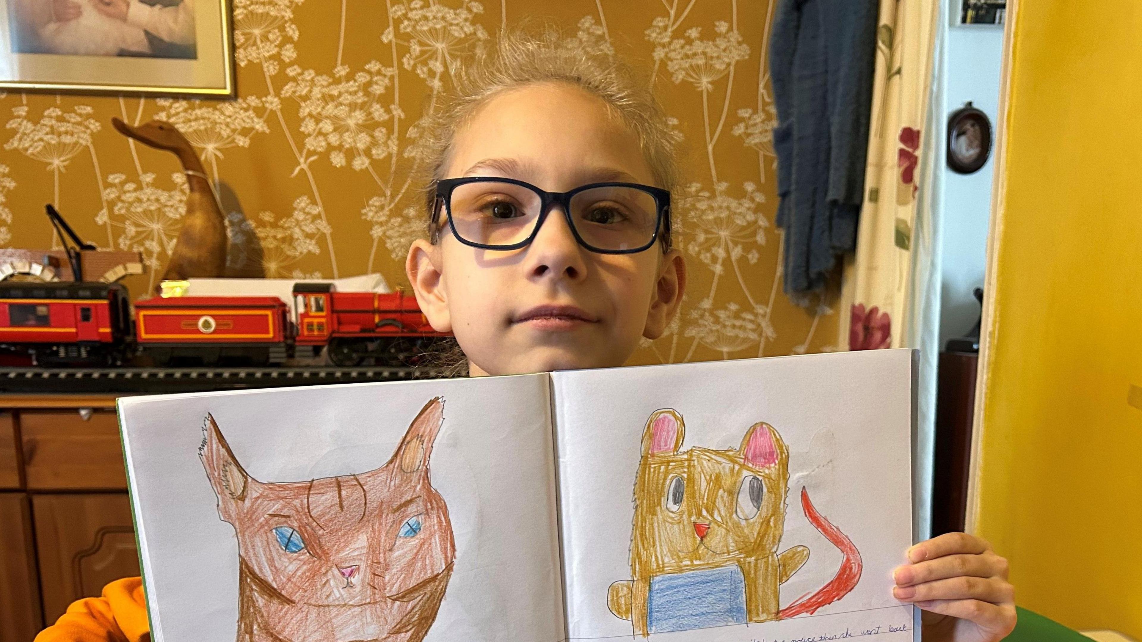 A white girl with black-framed glasses holds up a workbook. She has drawn a brown cat on the left page and a yellow mouse with a red tail on the right page. In the background is a red model train set and there are yellow walls.