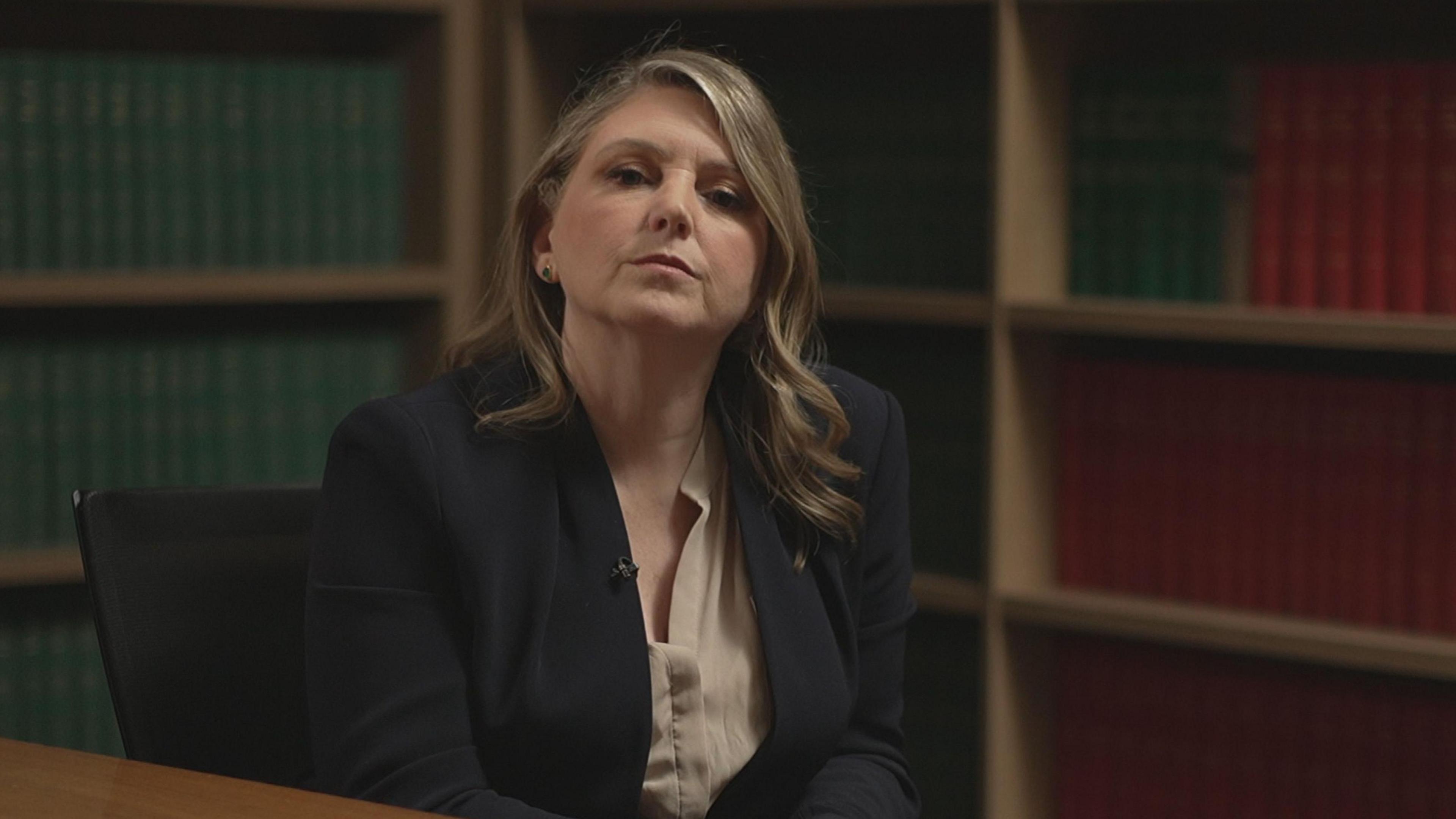 A woman with long, curly blonde hair looks towards the camera. She is sitting on a black chair in a formal room filled with red and green bound hardback books stacked on shelves. She is wearing a dark suit jacket and camel coloured blouse with one earring visible on her left ear. A small microphone is clipped to her suit jacket.