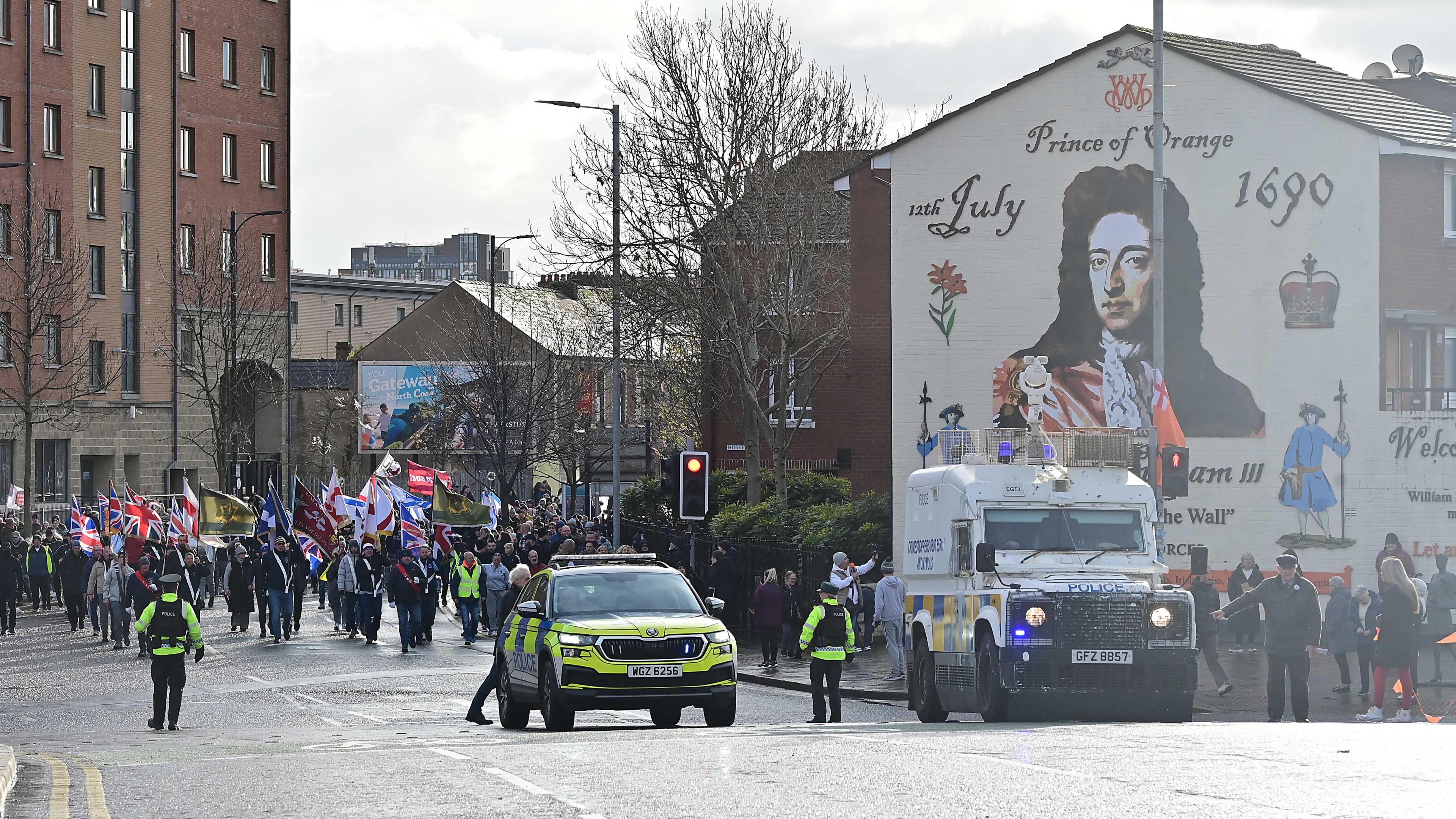 A PSNI car and van along with officers are in front of a procession of people who are waving a variety of flags. They are marching beneath a mural of King William III.