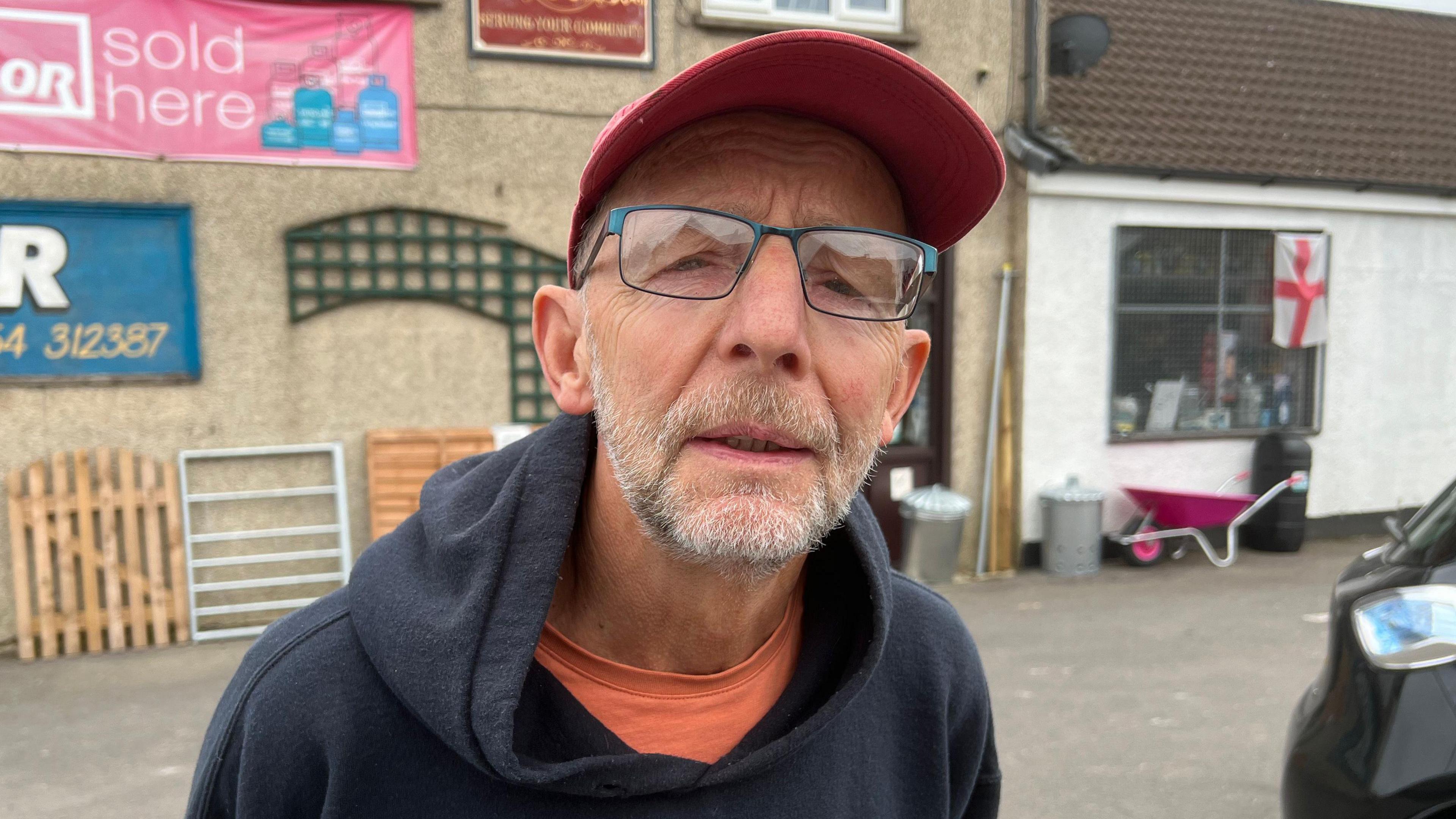 Martyn Page wears dark green glasses, a red hat, an orange top and a black hoodie. He stands outside a shop looking into the camera