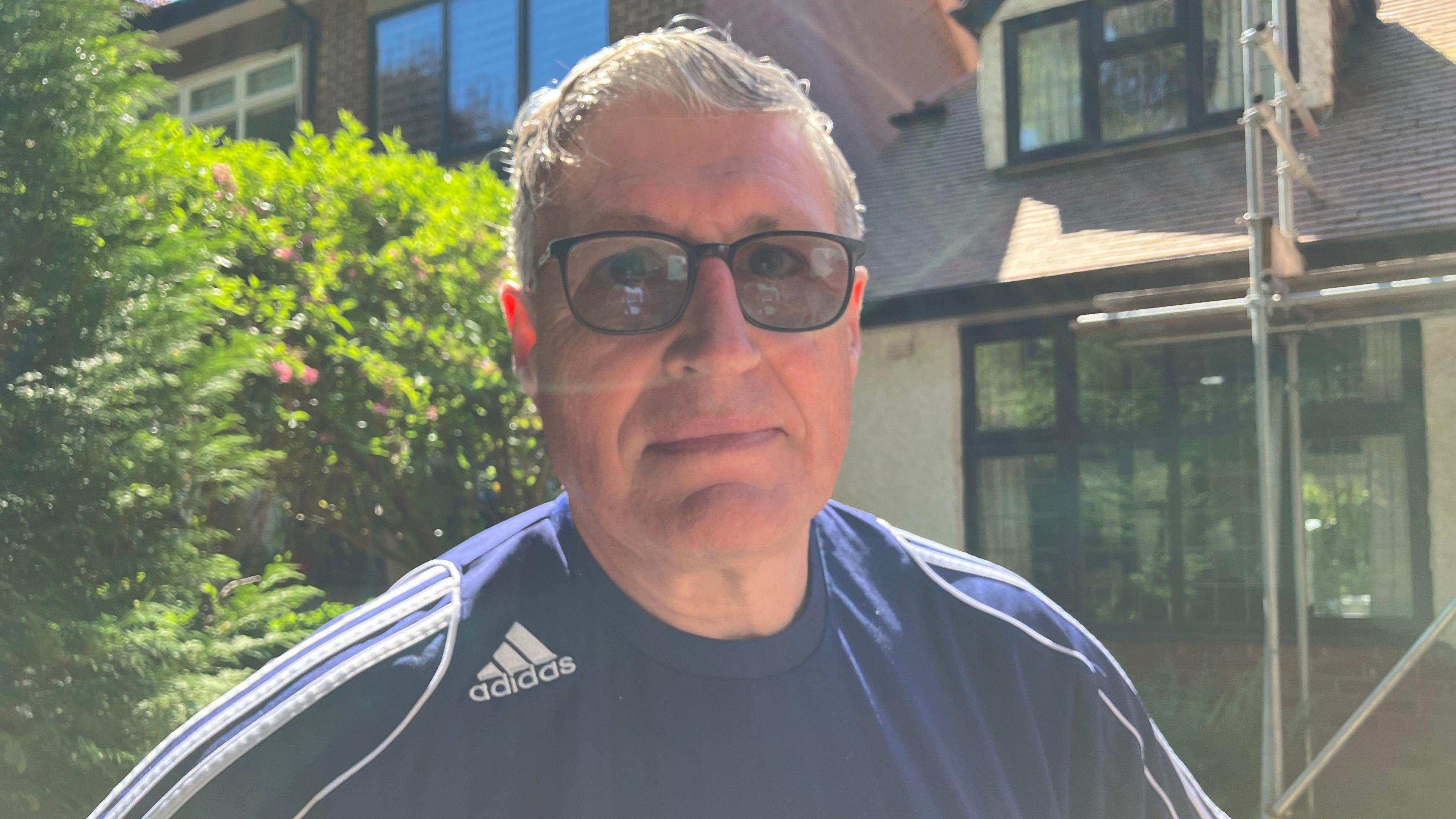 A man wearing a blue Adidas sports top with white stripes on both shoulders. He has grey hair and is wearing black tinted glasses. There is a house with scaffolding behind him.