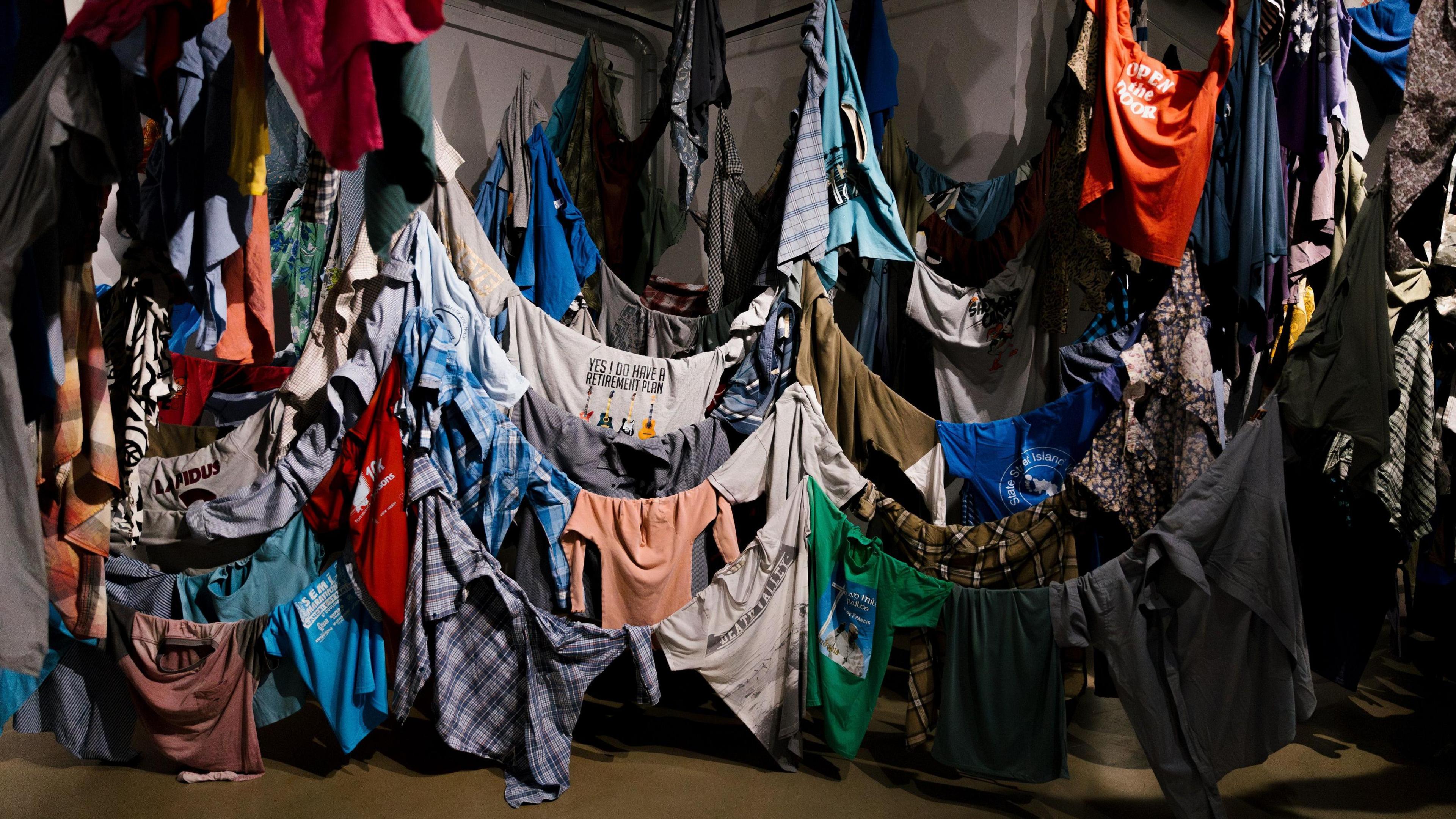 Shirts of different colours, designs and sizes are hung indoors around lines that drop down close to the floor where some of the shirts are touching the floor. There are hundreds of shirts on display.