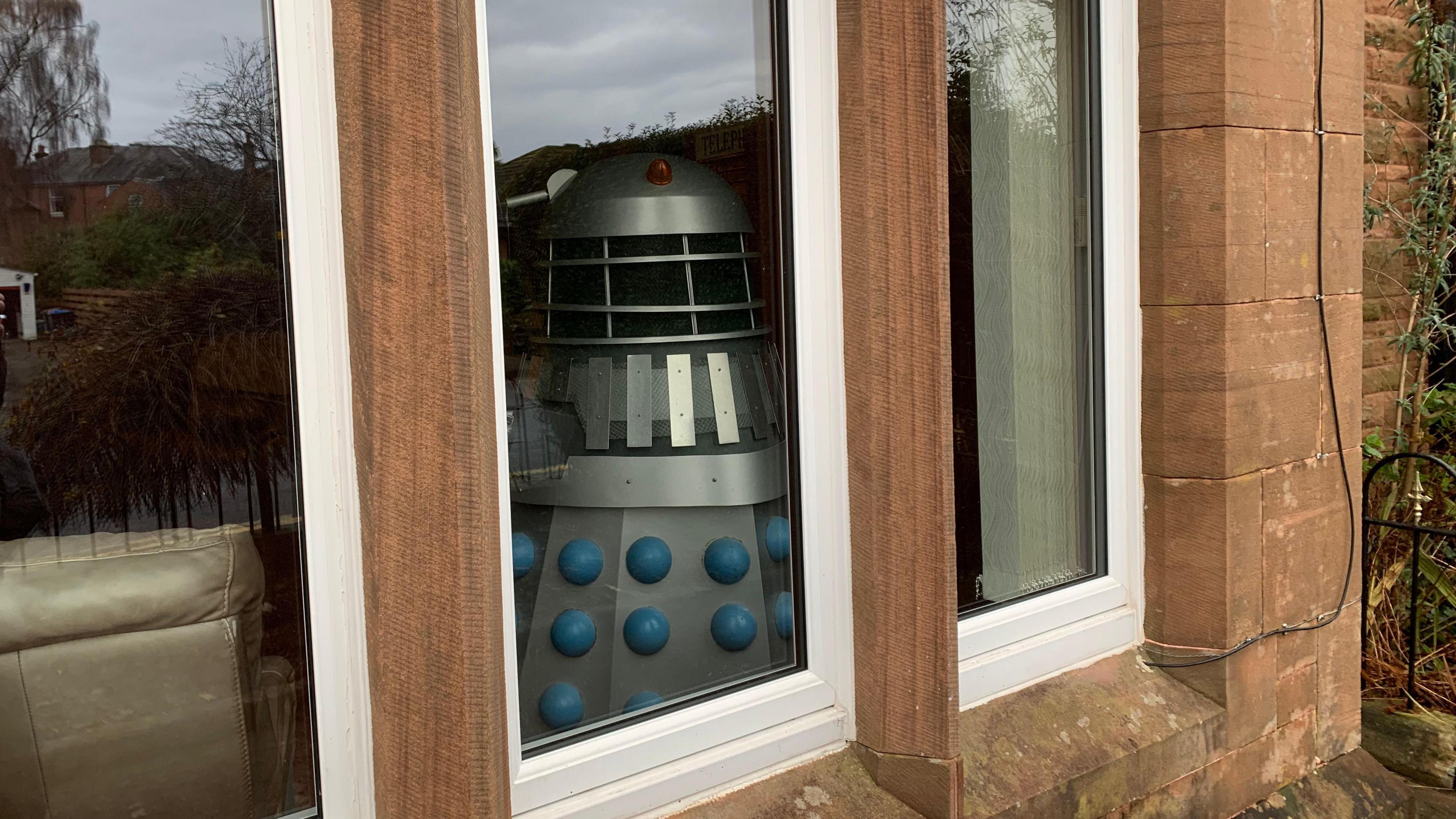 A domestic home window on a sandstone house with a Dalek visible in the living room