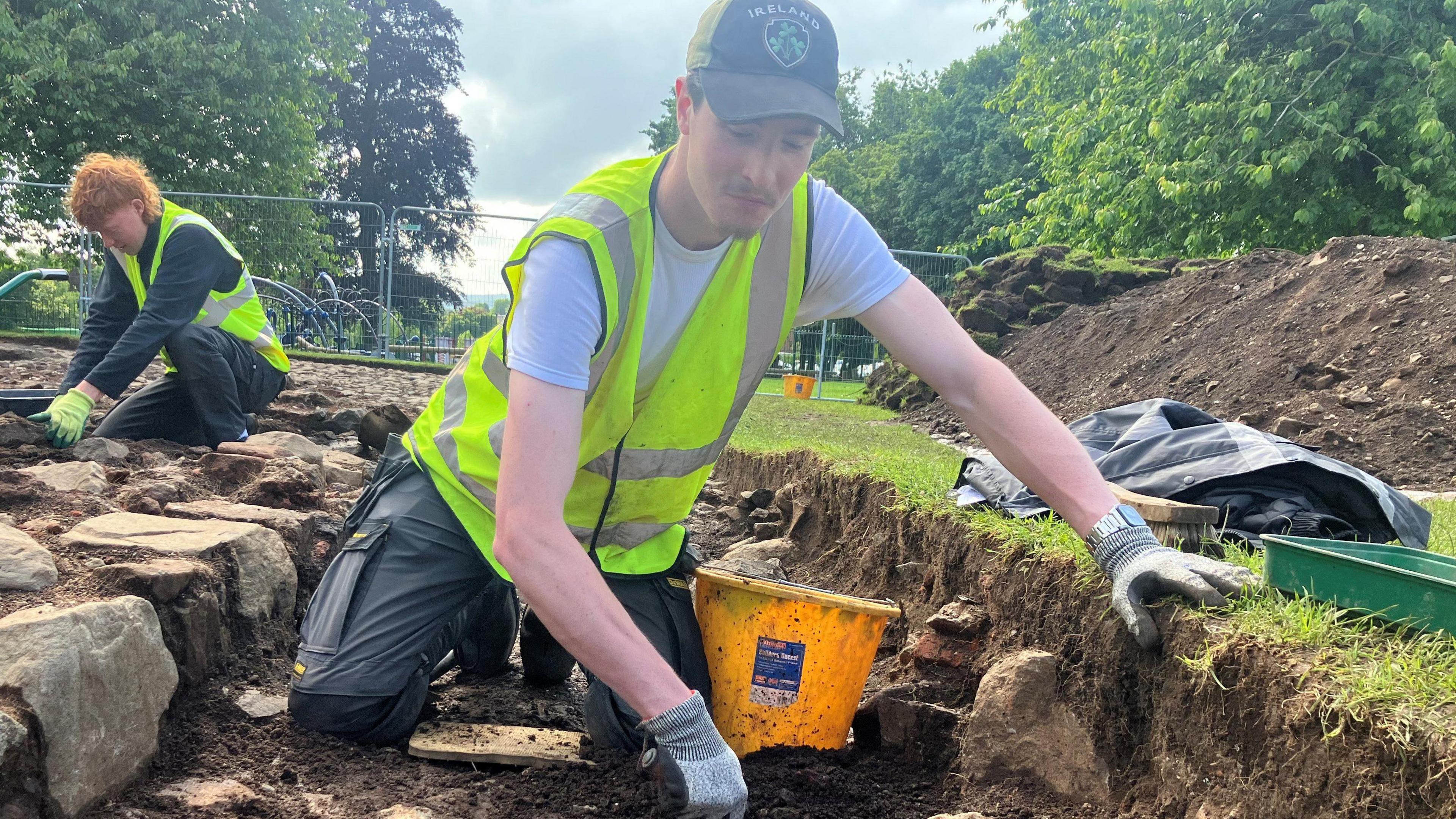 Arthur Redfern is digging in the soil. He is wearing a white t shirt, high visibility vest and cap. He also has grey gloves on. He is shovelling into a yellow bucket. In the background is another person, a grey fence and some large trees on green grass.