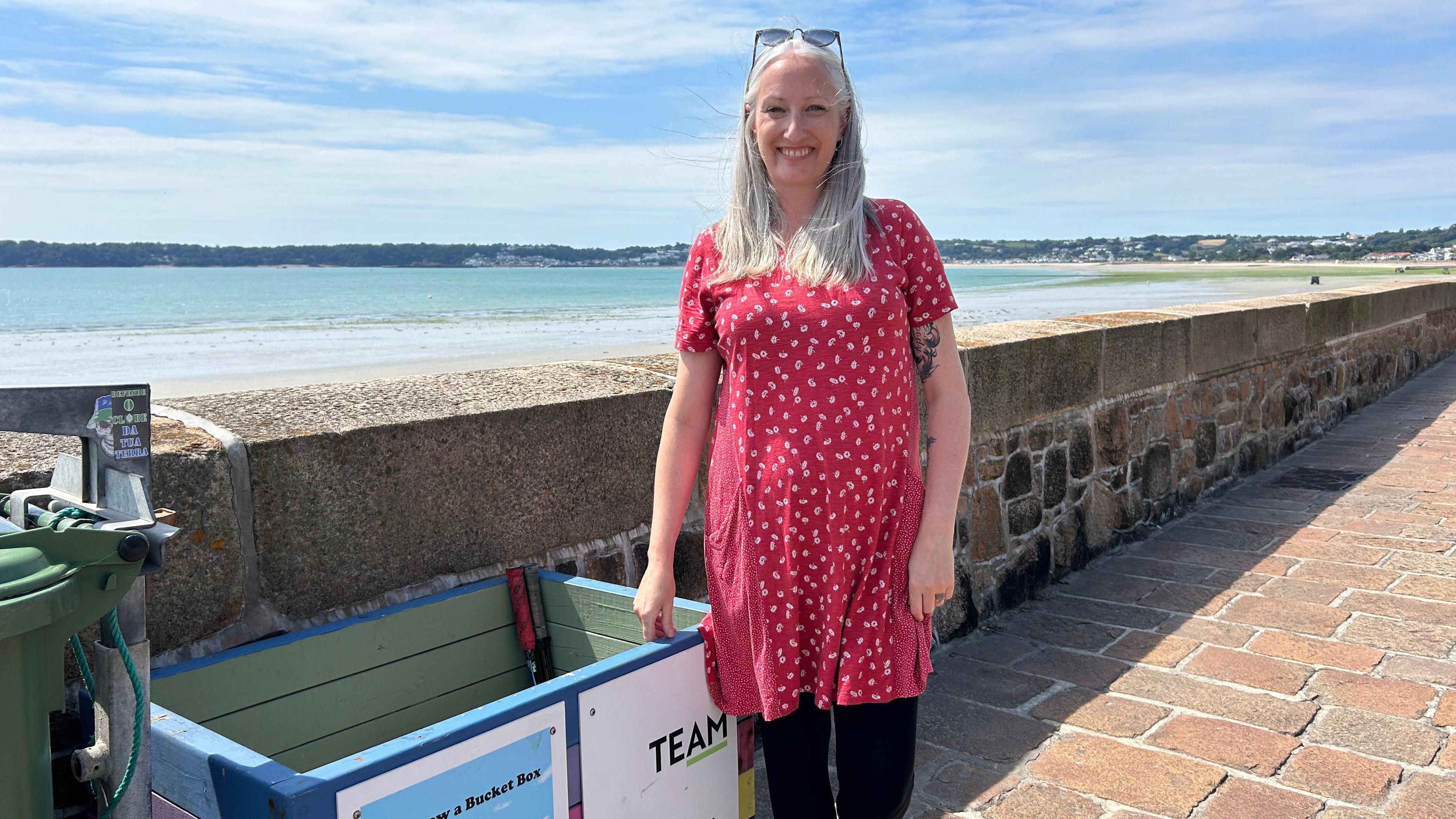 Louise Carson is wearing a red dress and stood on the promenade by St Aubin's Bay.