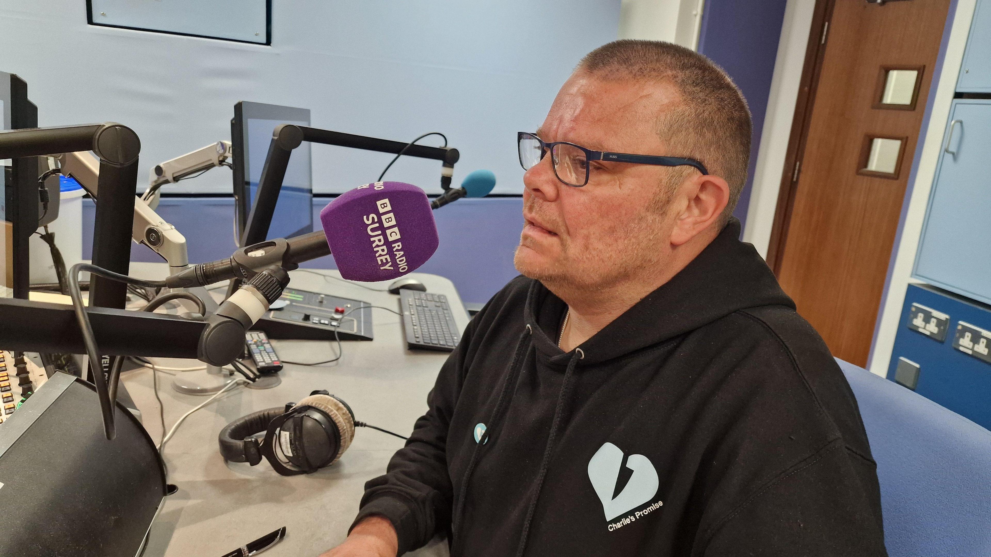 Martin Cosser sitting in the BBC Radio Surrey studio, wearing a black polo neck shirt.