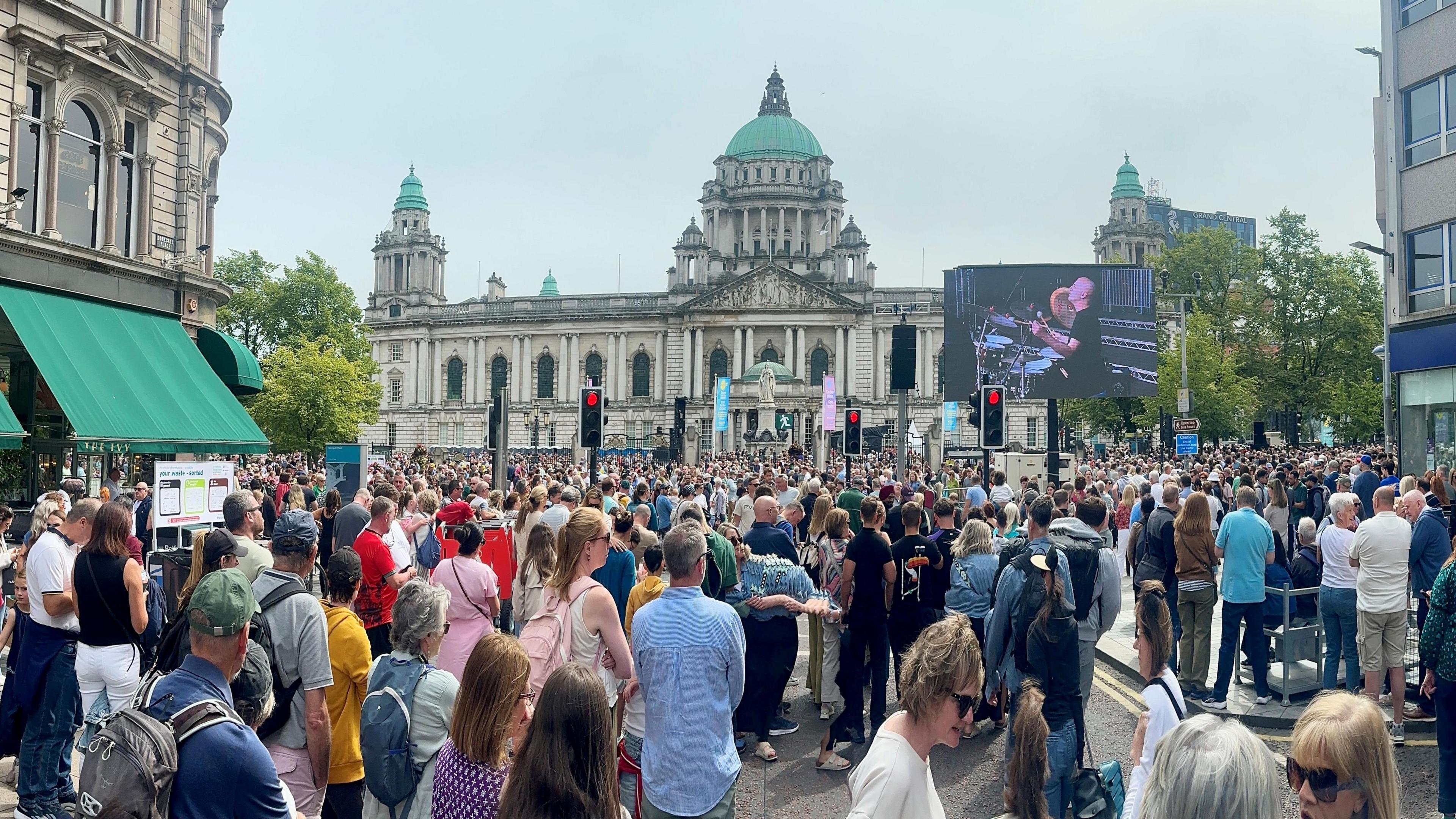 A large crowd in front of Belfast City Hall - it is a white Baroque style building with a green dome. There is a large TV screen on the right of the picture