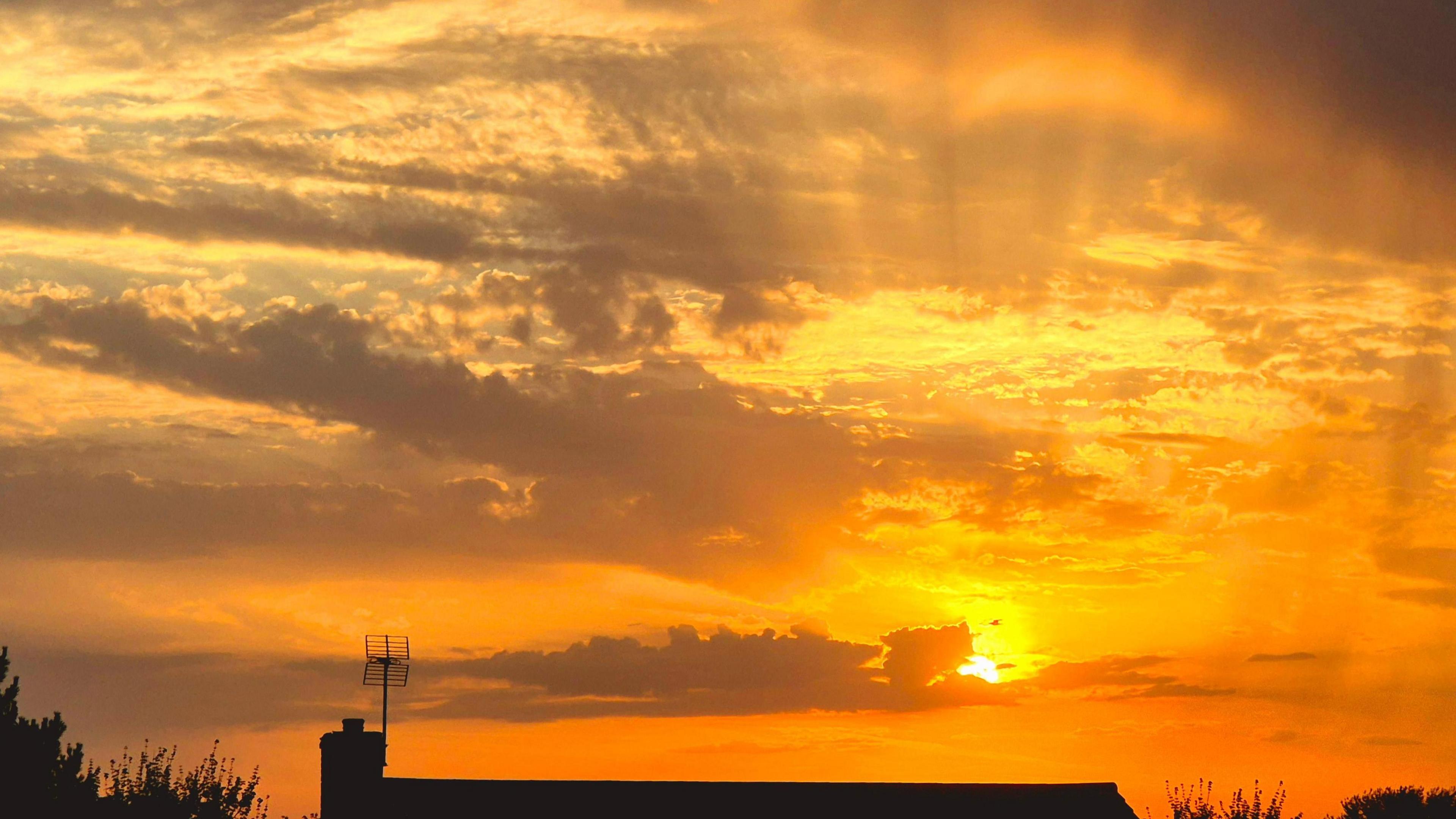 Silhouette of the top of a house with a traditional ariel on top.  Cloud fills the sky but turned yellow and orange by the sunset with sunbeams coming from the partially blocked sun behind a cloud.