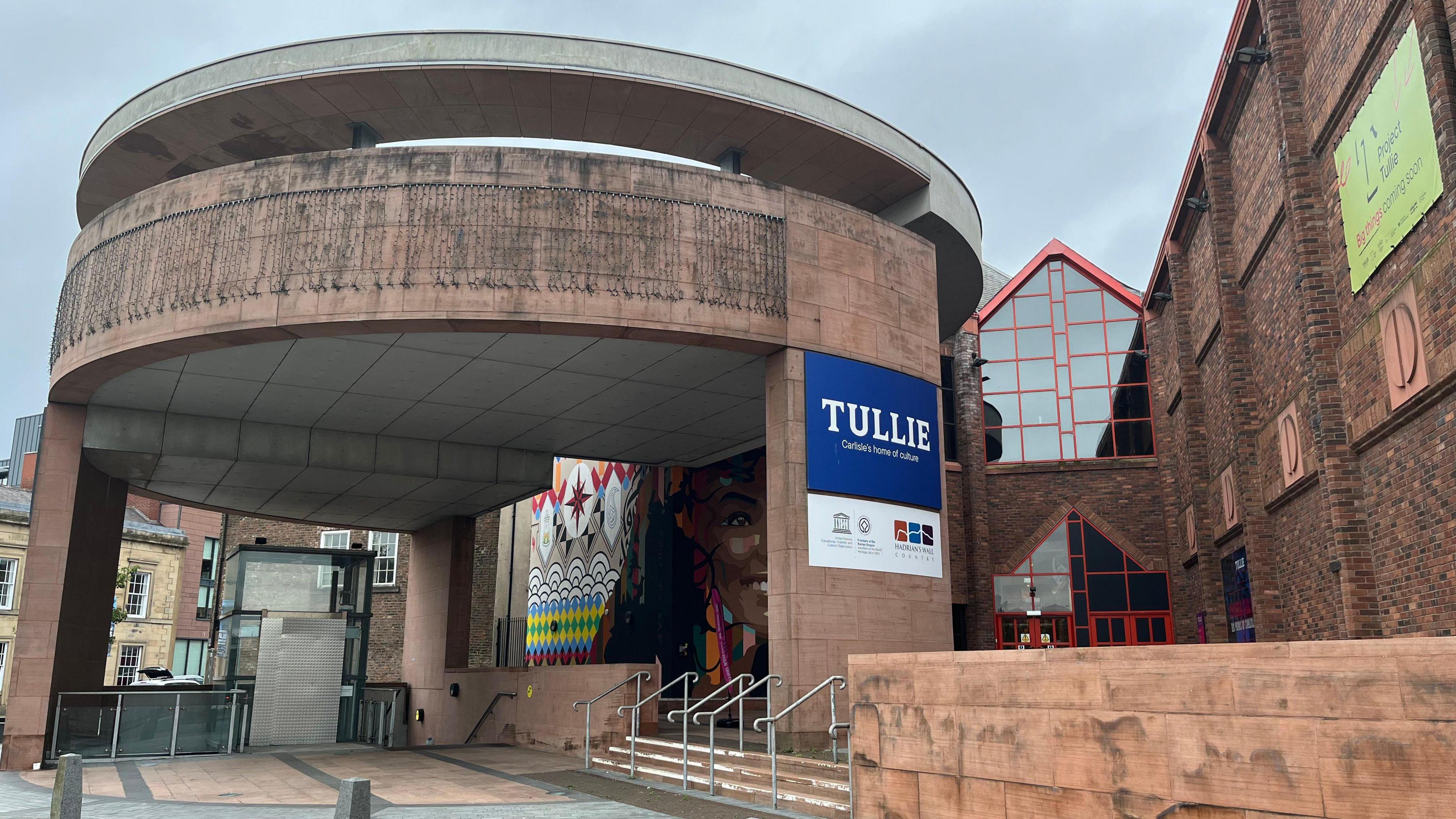 A general view of the entrance to Tullie in Carlisle. The entrance of the museum is formed of a circular building with a colourful mural in the background.