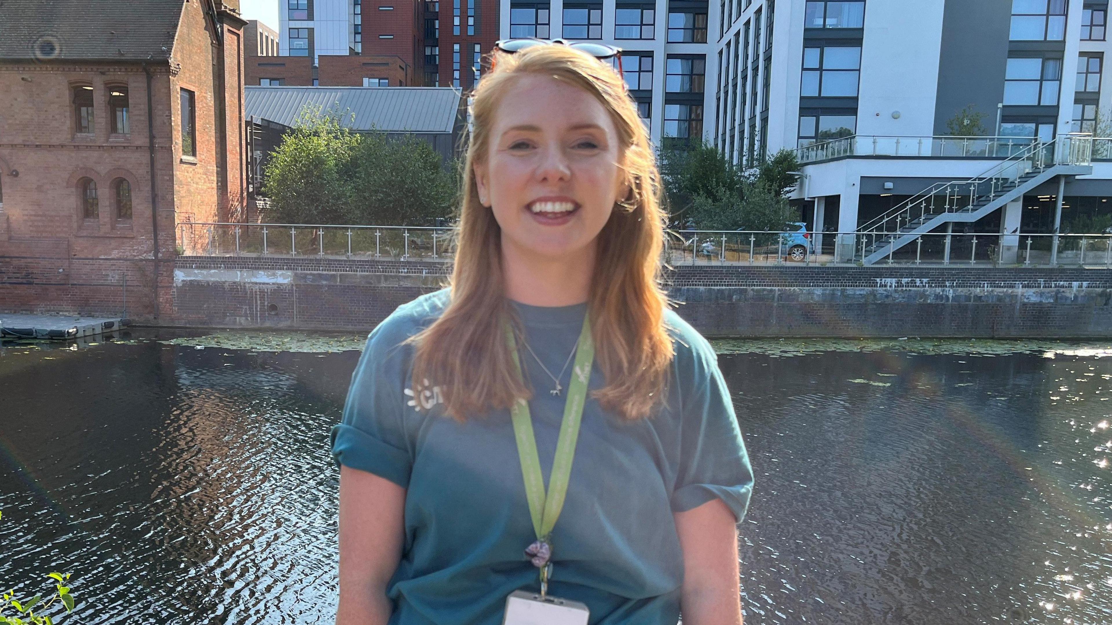 Juliette Burt smiling at the camera wearing a turquoise top and a lanyard. She is stood in front of the river