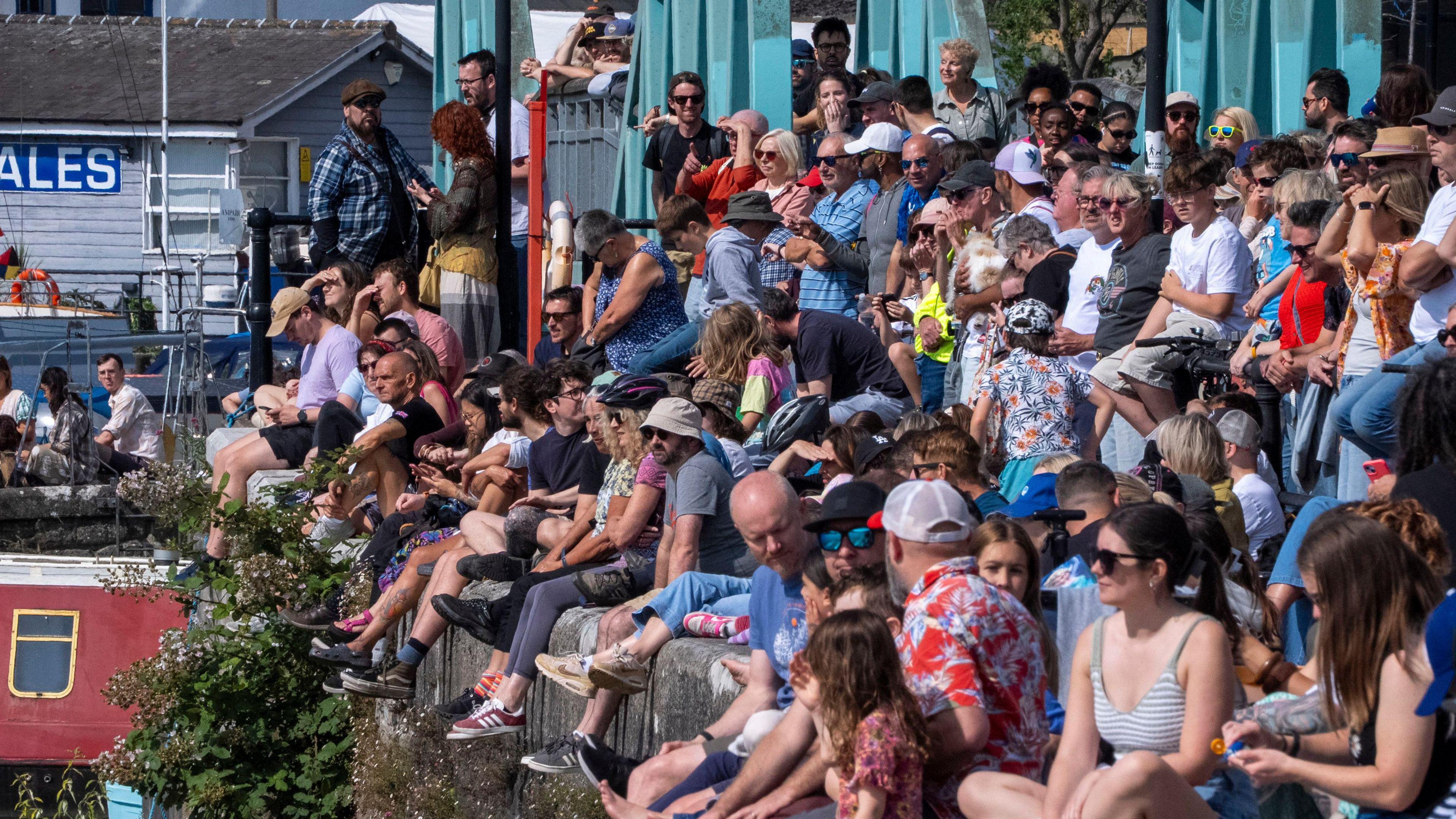 People sitting on the edge of the harbour wall, with more standing behind.