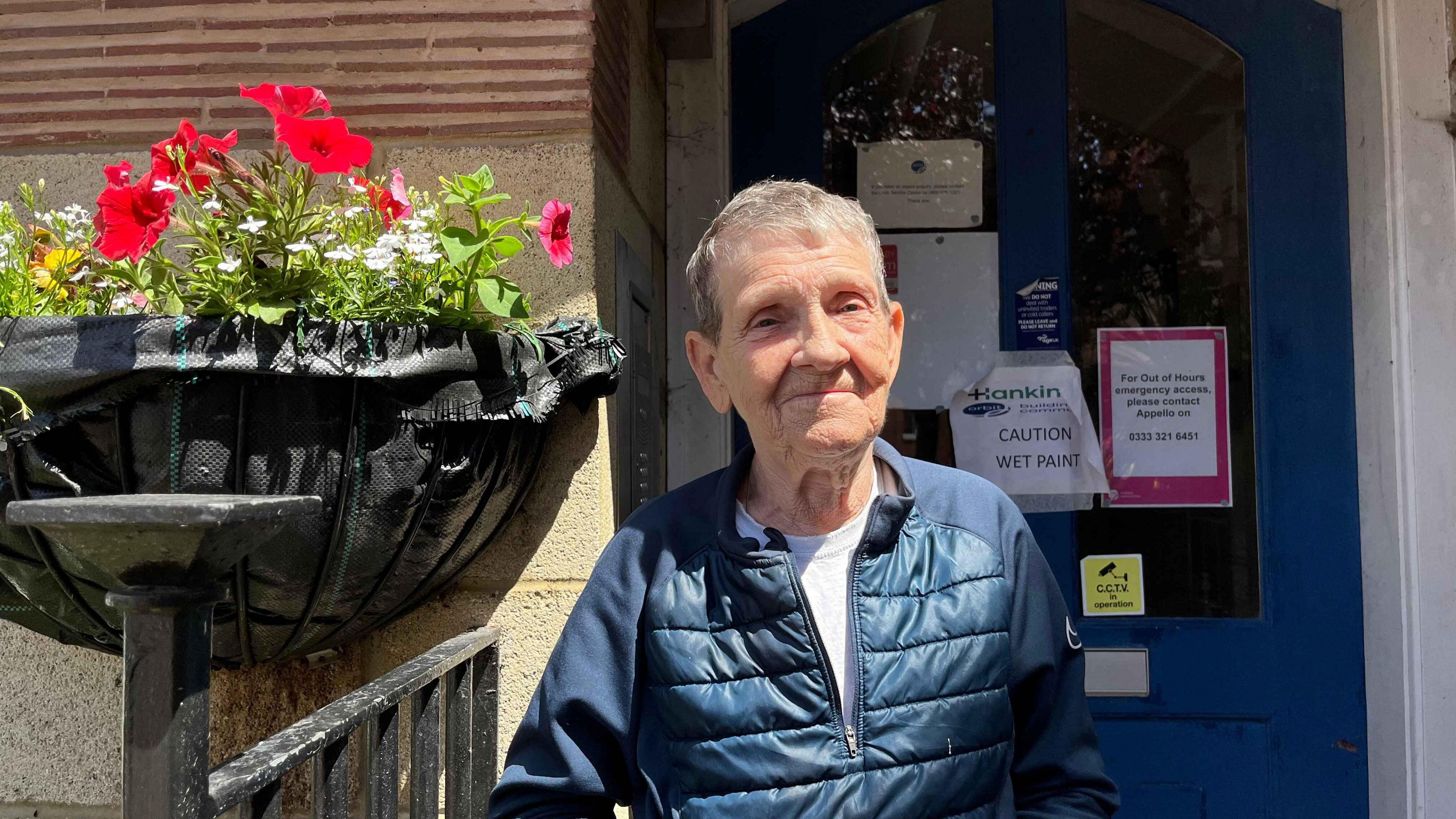 Chris Parrish wearing a jacket outside the front door of the flat which is blue. Next to her is a hanging basket of flowers