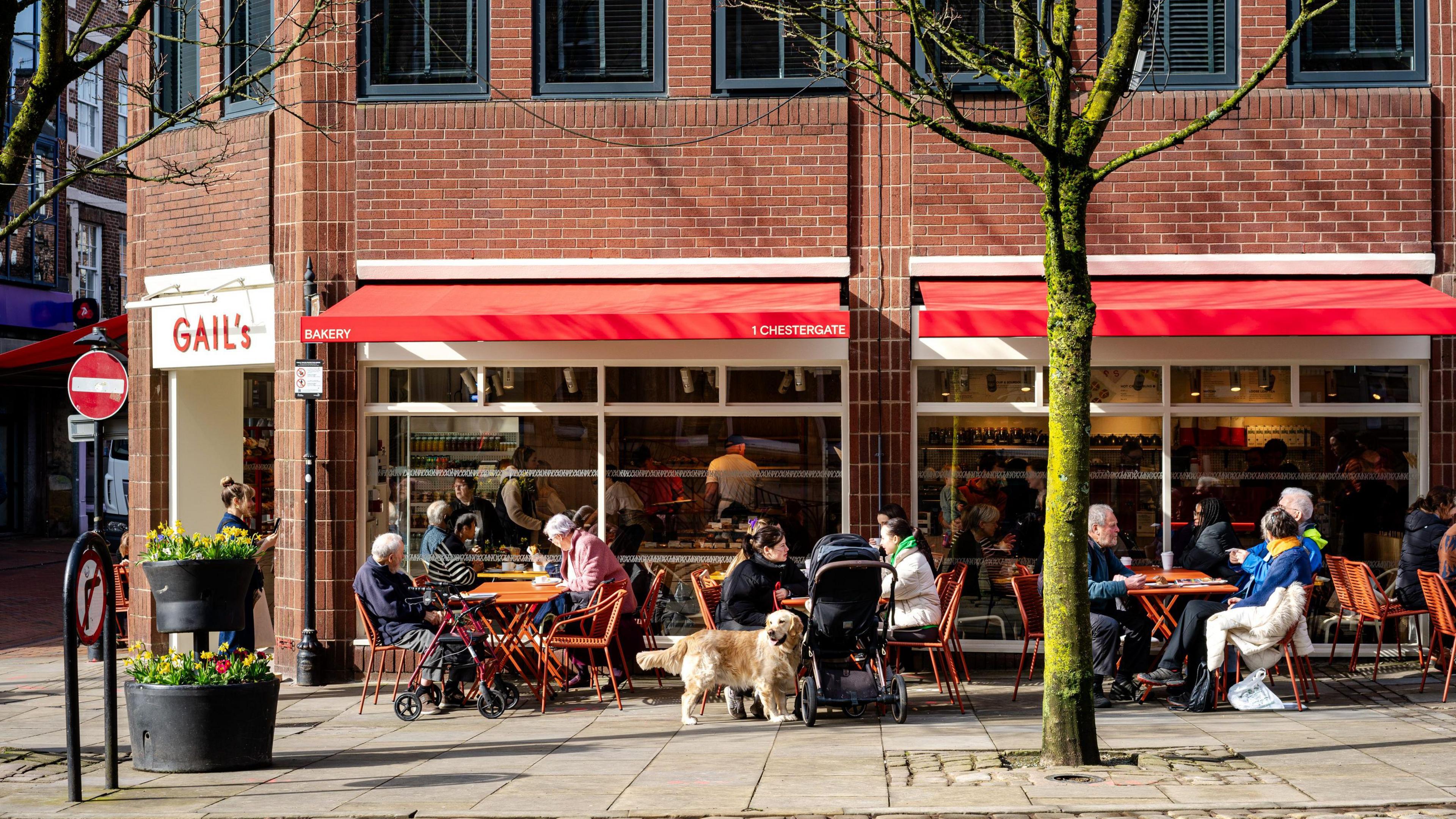 People sitting at a table outside Gail's cafe, one with a golden retriever and a buggy