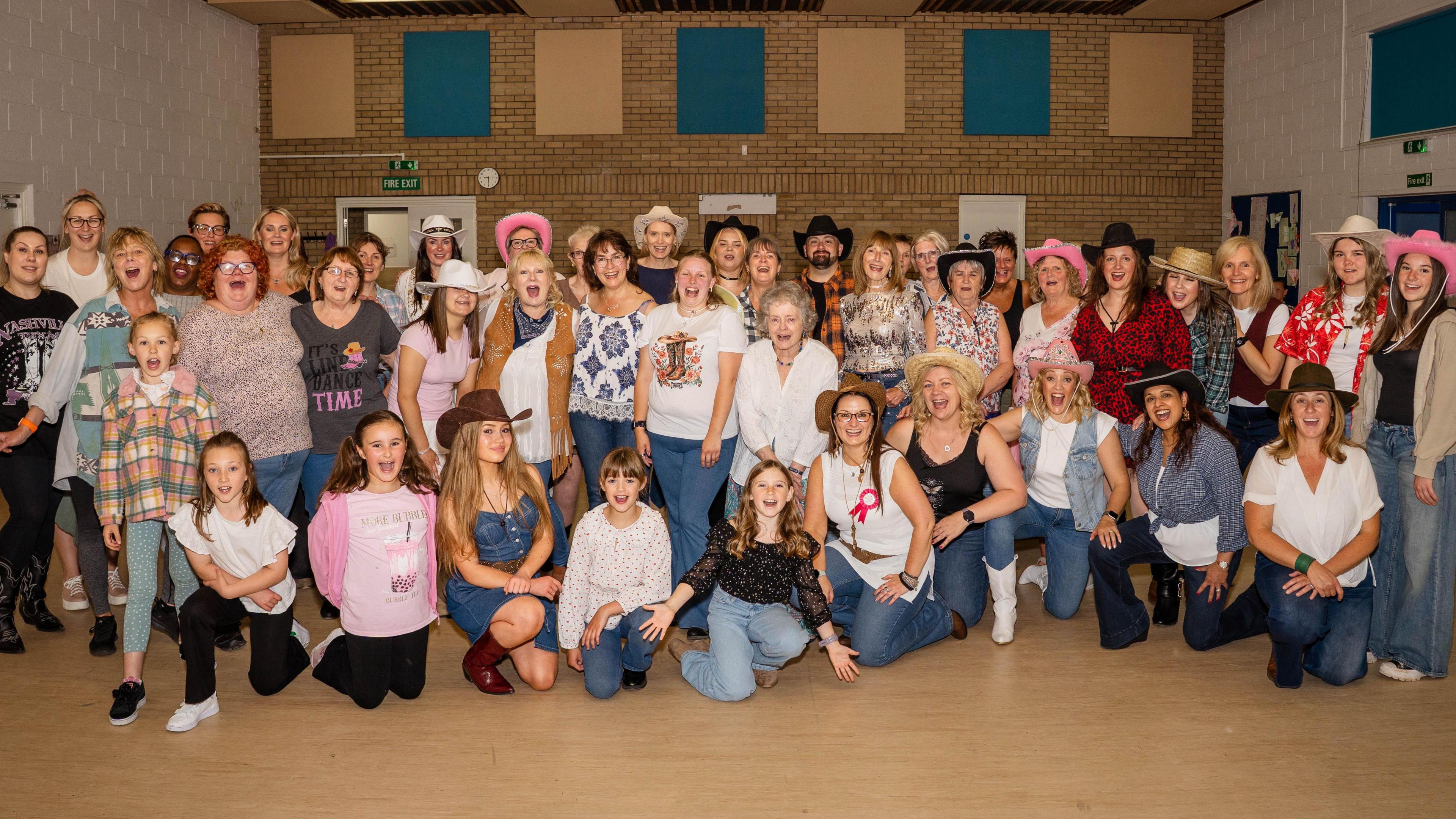 A large group of people, mostly female, of a range of ages stood and crouched in rows, facing the camera. many of them are wearing cowboy hats, denim and check shirts.