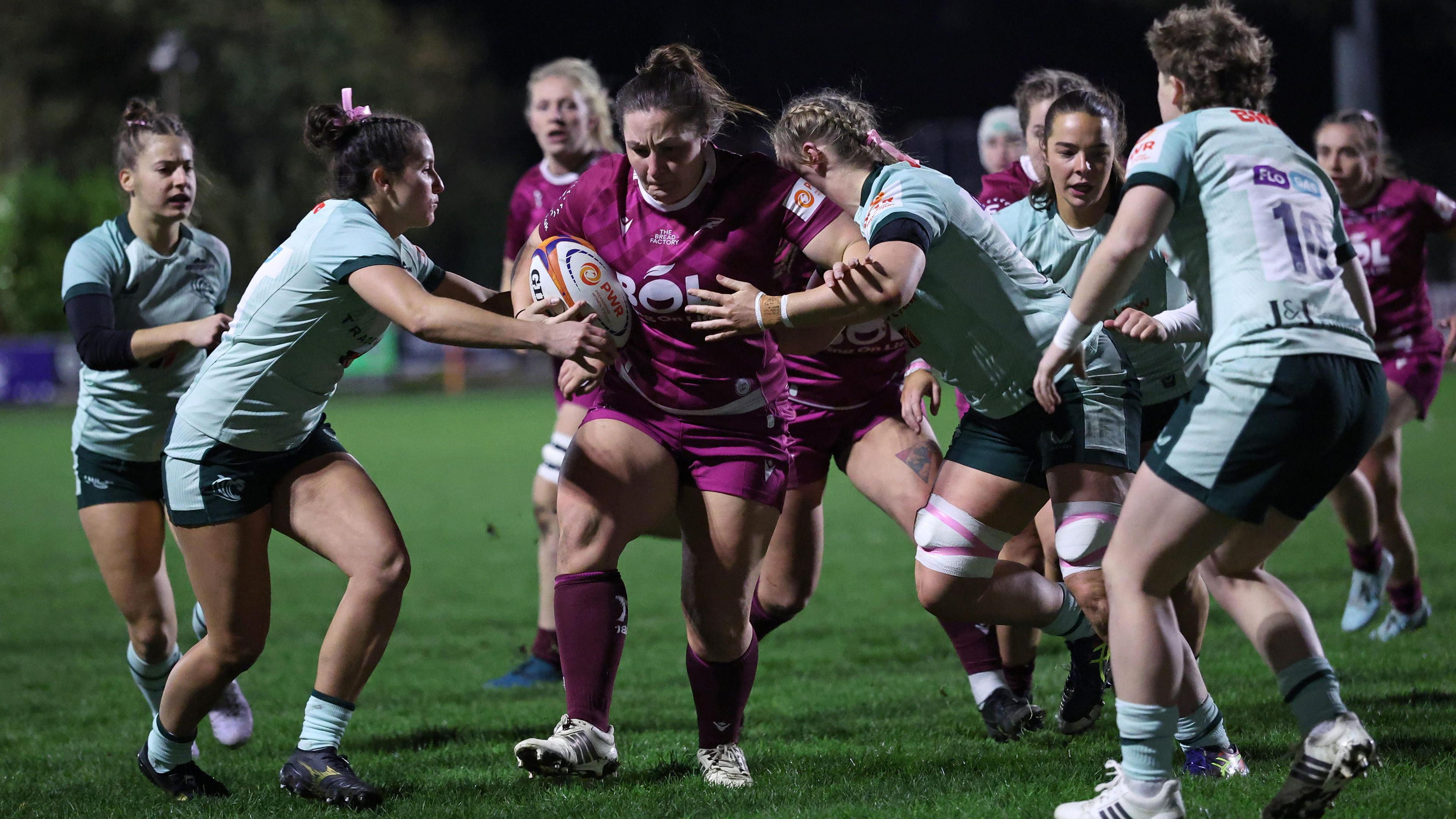 A photo of Amy Cokayne making a run for rugby football union club Sale Sharks on a grass pitch in a Premiership Women's Rugby match against Leicester Tigers. Cokayne is wearing a purple kit and holding a white, orange and blue ball while being challenged by Leicester players in light green shirts and socks with light green and dark blue shorts. Some of Cokayne's teammates are visible in the background
