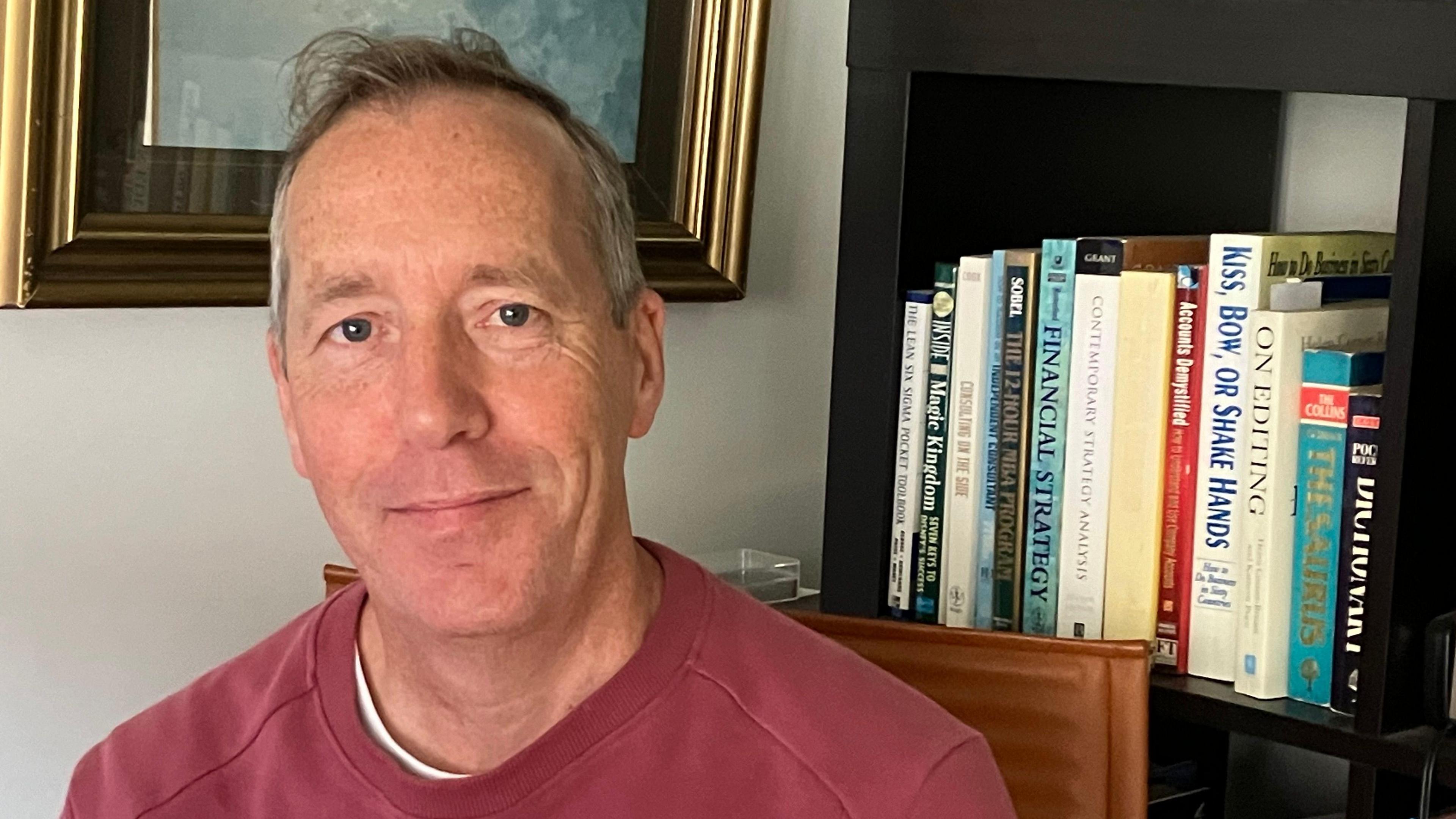 Simon Eyre wearing a red top sits in front of a bookcase