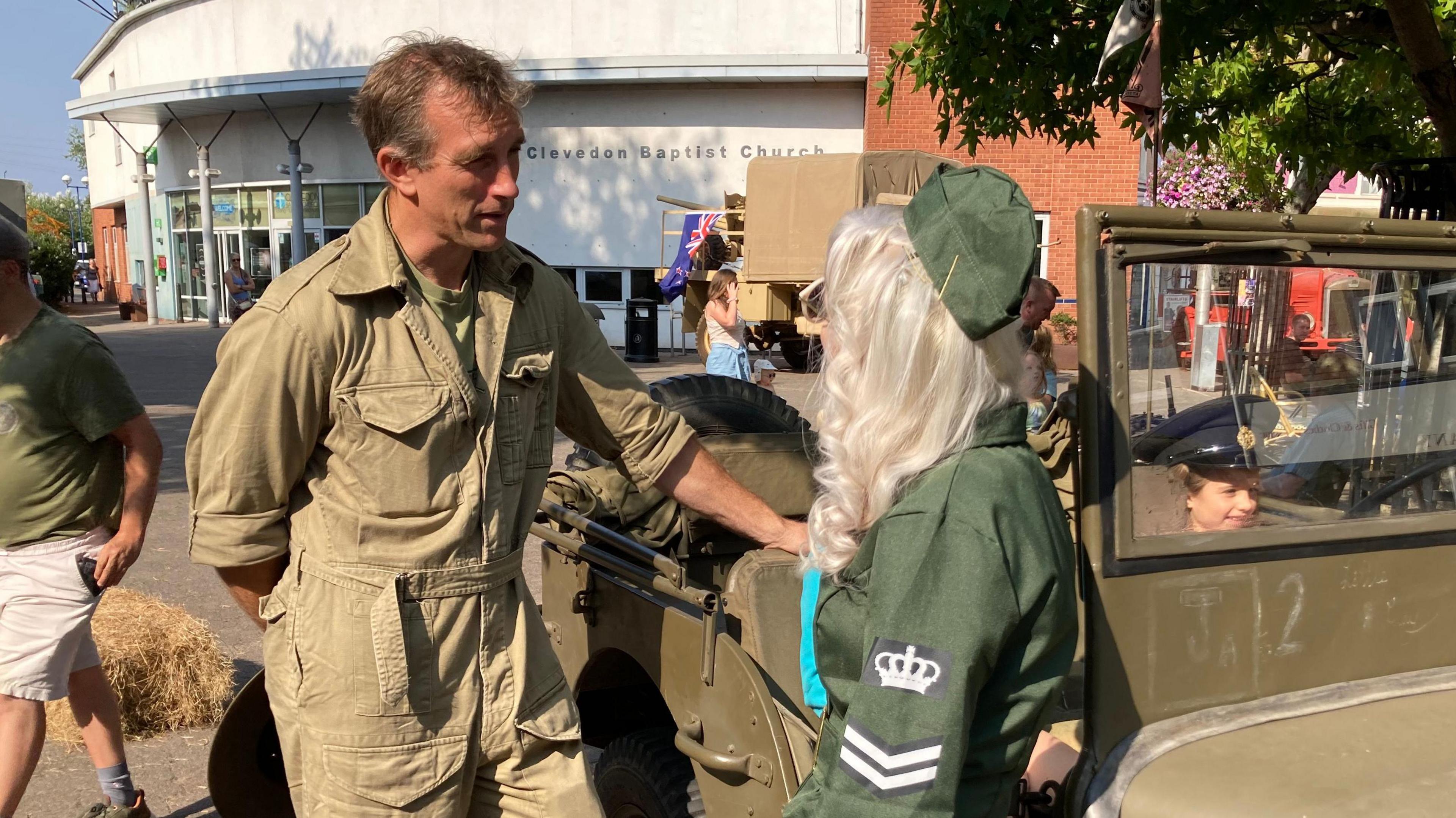 James leans with one hand on a green military vehicle, wearing a green khaki boilersuit. He is speaking to a woman with long bright blonde hair. who is wearing green 1940s costume. 