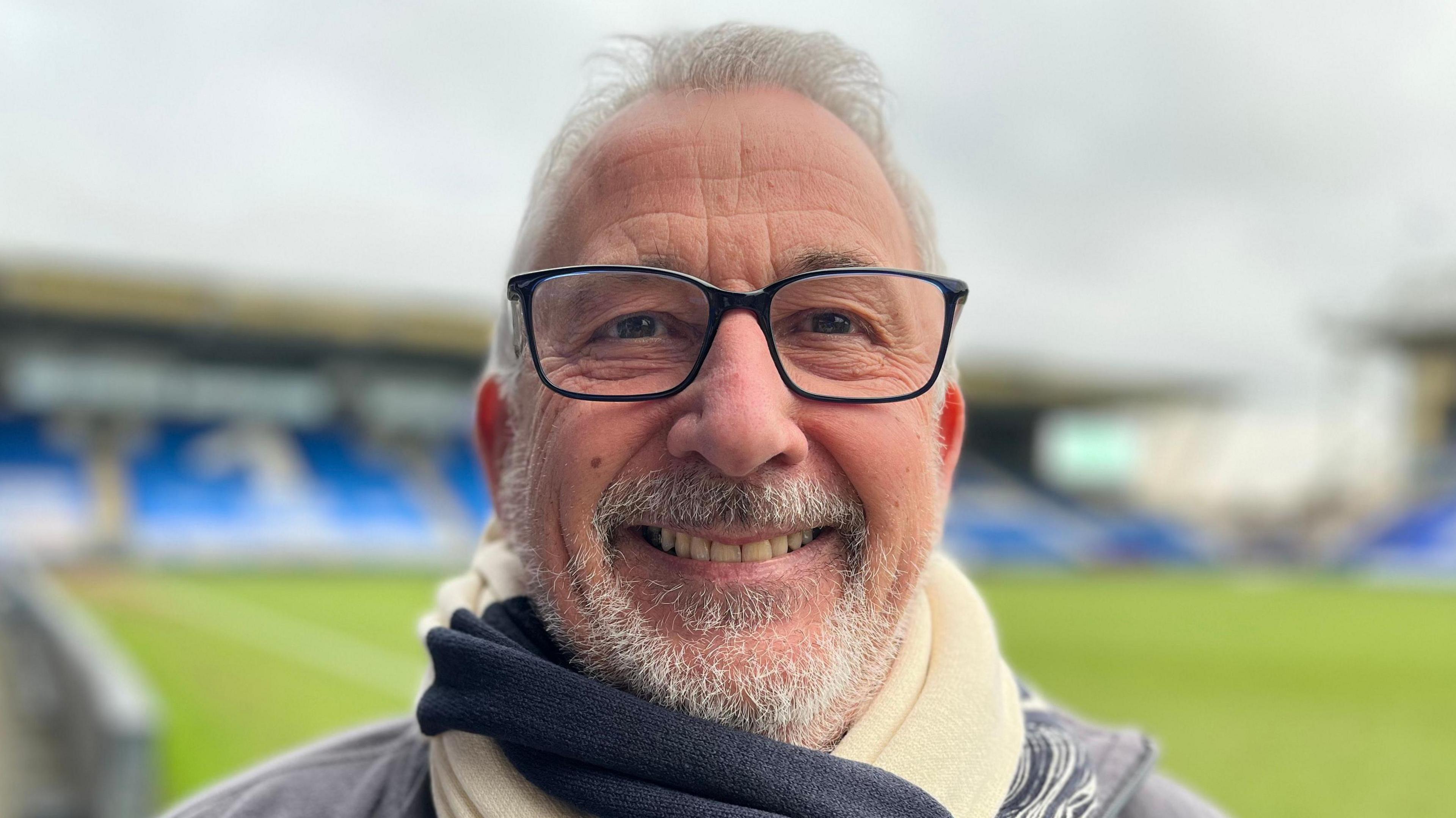 Michael Ferguson is smiling at the camera while stood inside the Weston Homes Stadium. The pitch behind him is empty, as are the blue seats in the stands. Michael has grey facial hair and grey hair as well as black square framed glasses.  He has a blue and white scarf wrapped around his neck. 